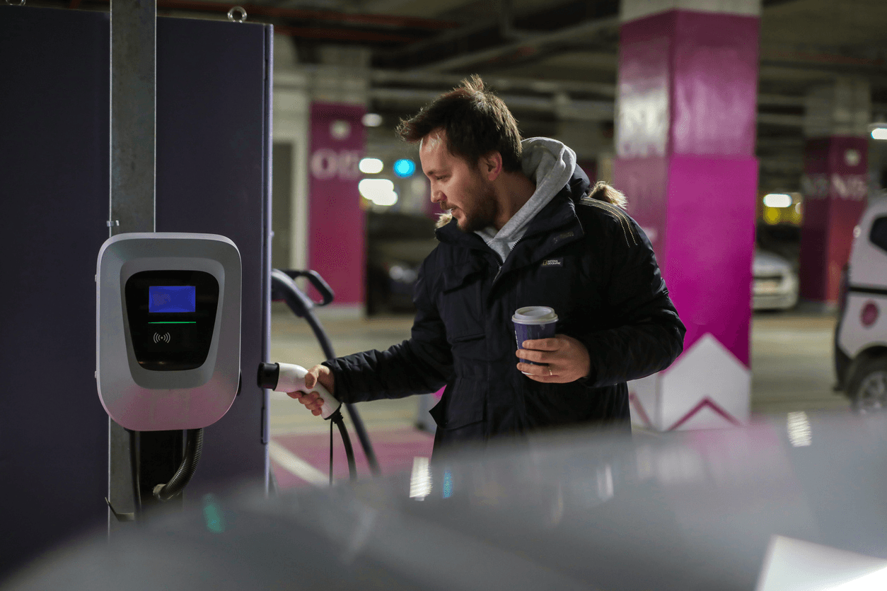 Man in a coat and hoodie holding a coffee charging his car in an underground carpark