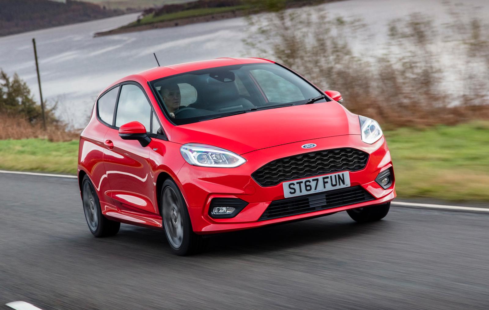 a red Ford Fiesta hatchback driving on a country road alongside a lake