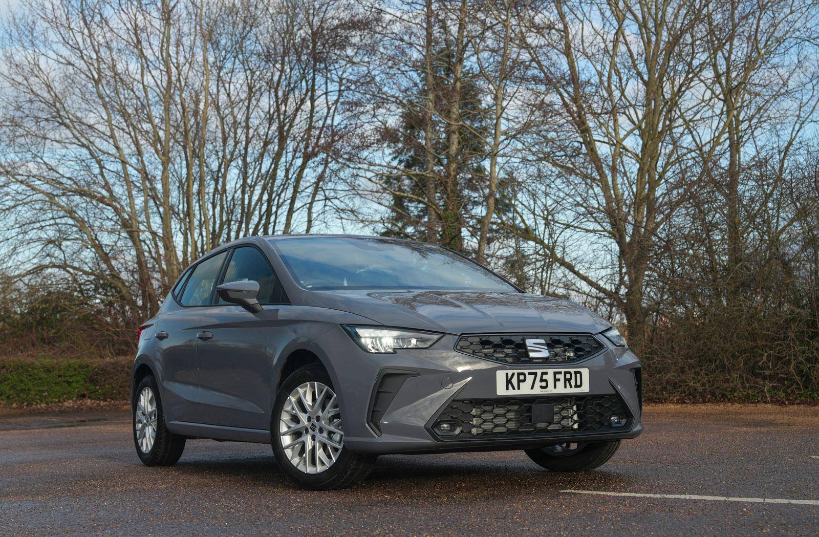 A grey hatchback car parked on a wet road, with trees in the background under an overcast sky.