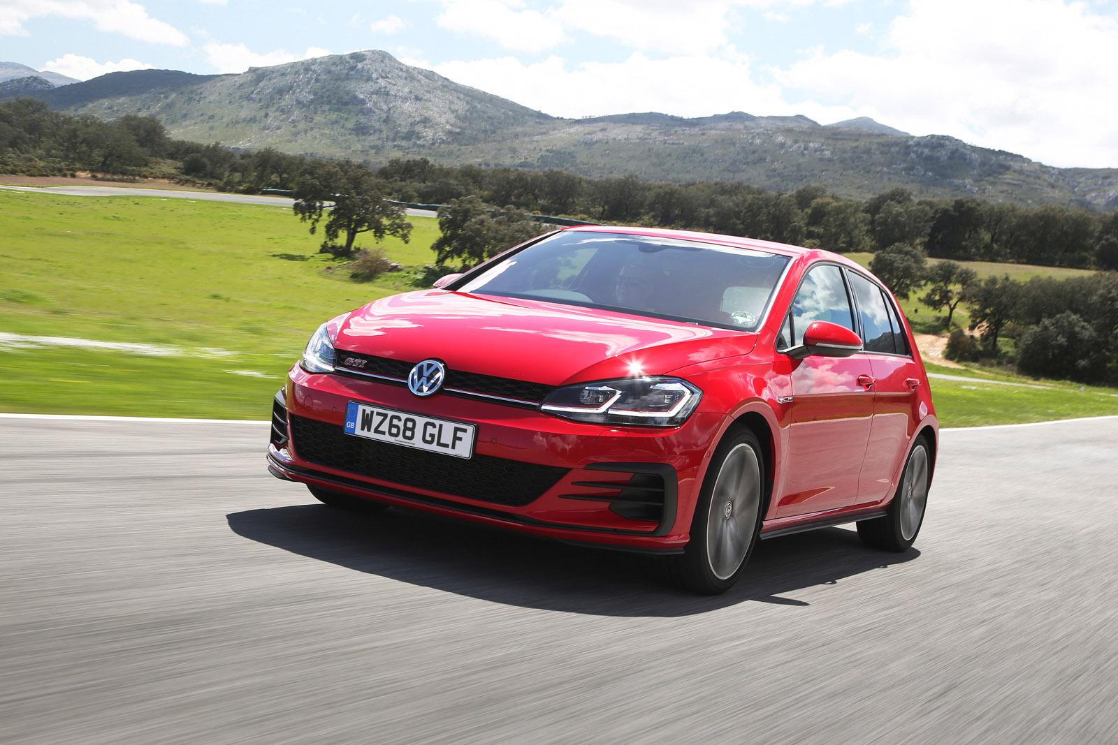 Red Volkswagen Golf GTI driving on a scenic road with mountains and green fields in the background.