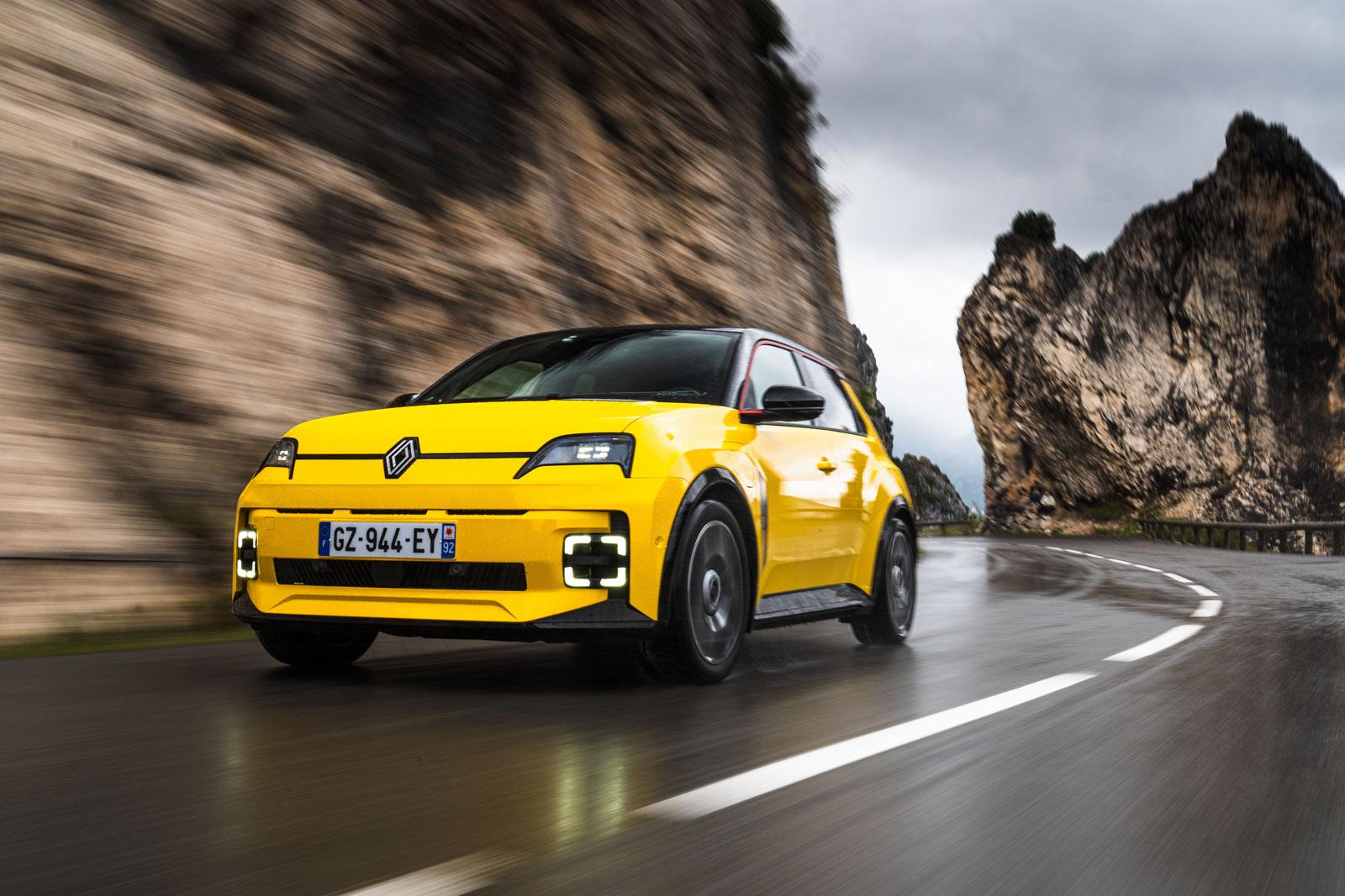 Yellow car driving on a wet, winding mountain road with rocky cliffs and a cloudy sky in the background.