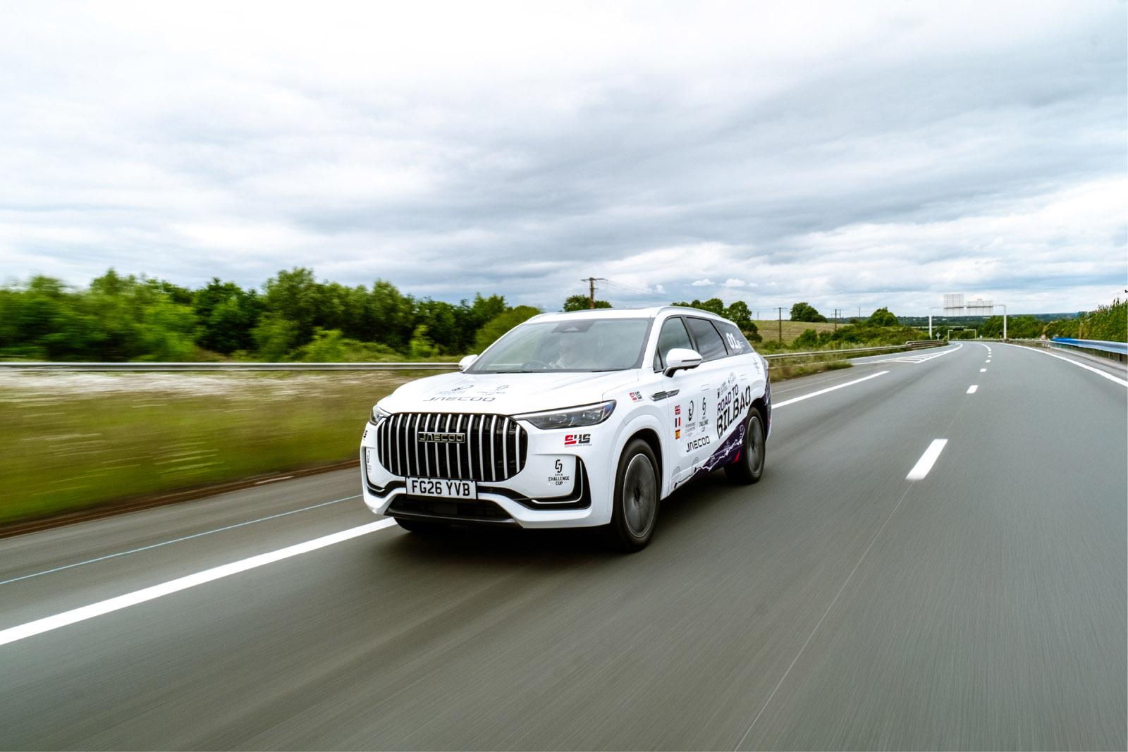White SUV covered in sponsor logos driving on a highway with greenery and cloudy skies in the background.