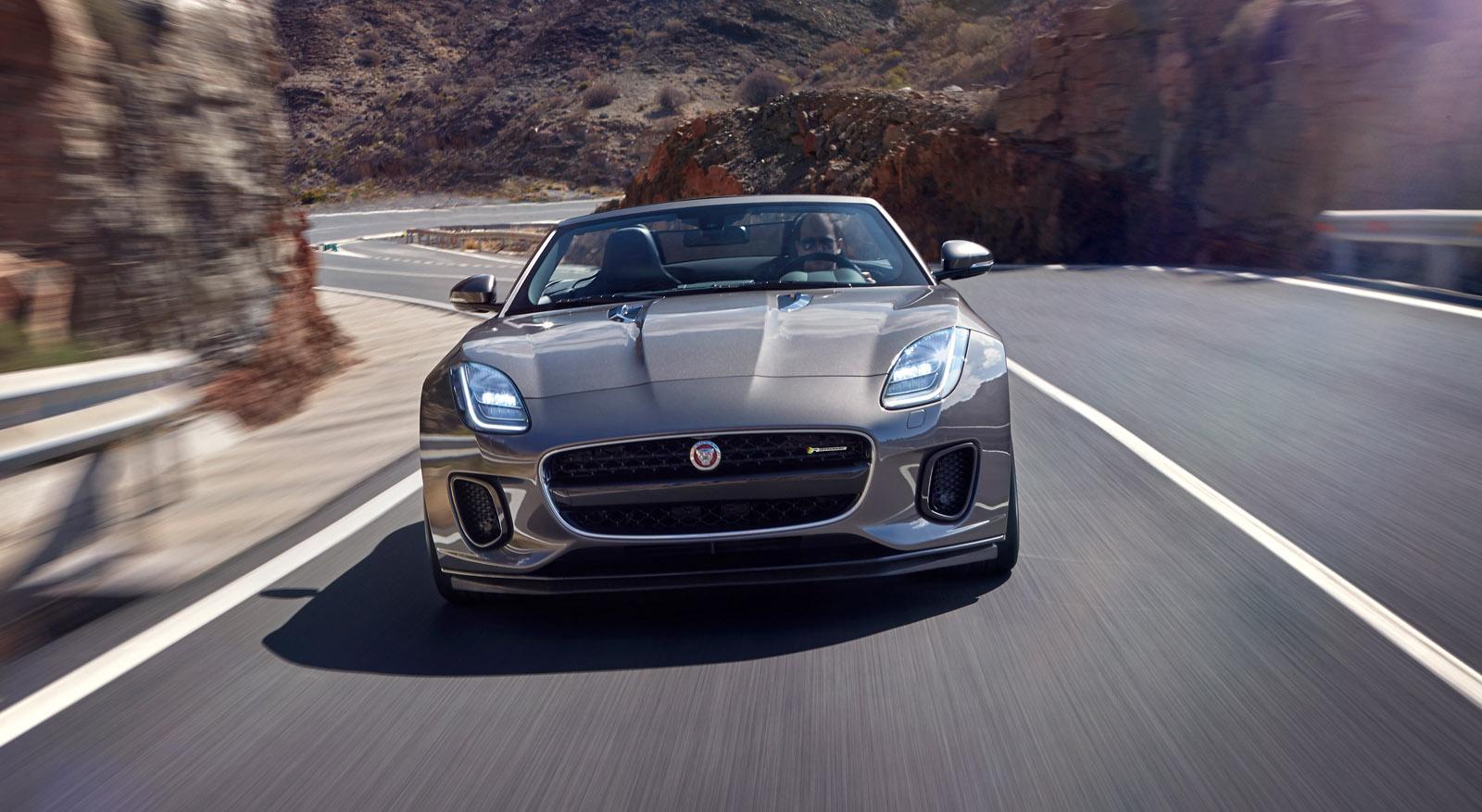 Silver sports car driving on a winding mountain road with rocky terrain and clear skies in the background.