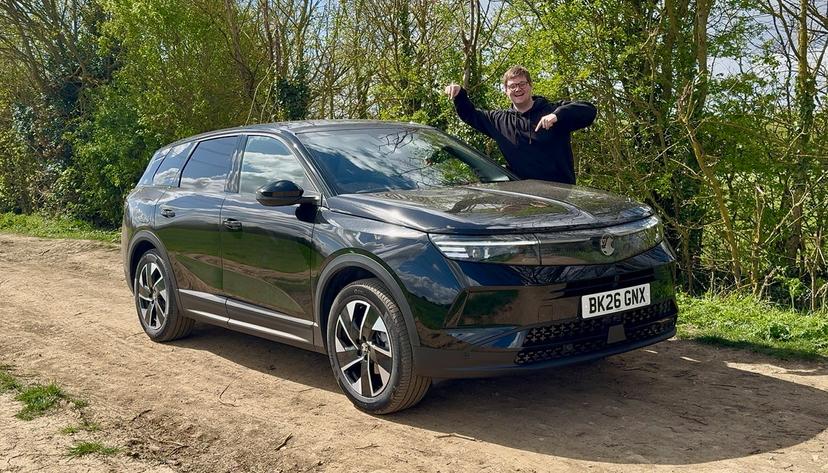A person in a black hoodie excitedly points at a sleek black car parked on a dirt road, surrounded by greenery.