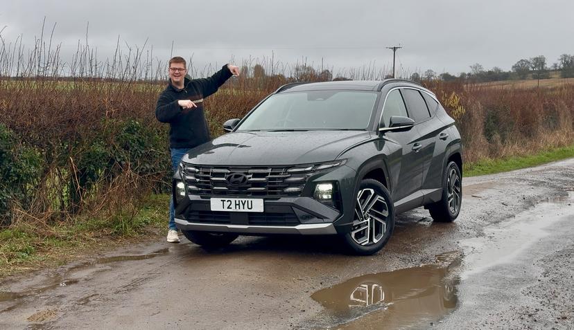 A person stands next to a grey Hyundai SUV on a wet rural road, smiling and pointing at the car. Overcast sky and bushes in the background.