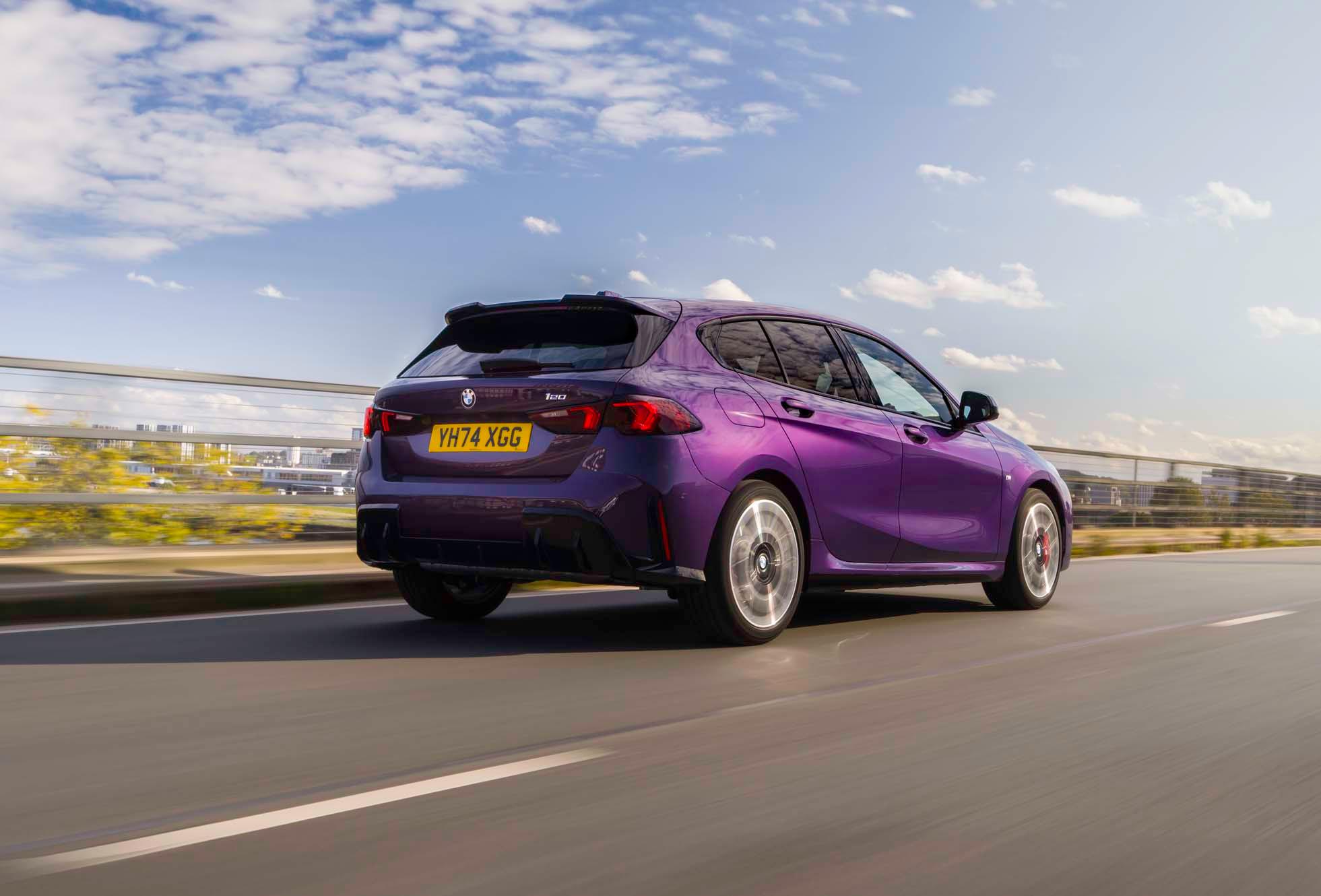 A purple hatchback car drives on a motorway under a clear blue sky, with cityscape and greenery in the background.