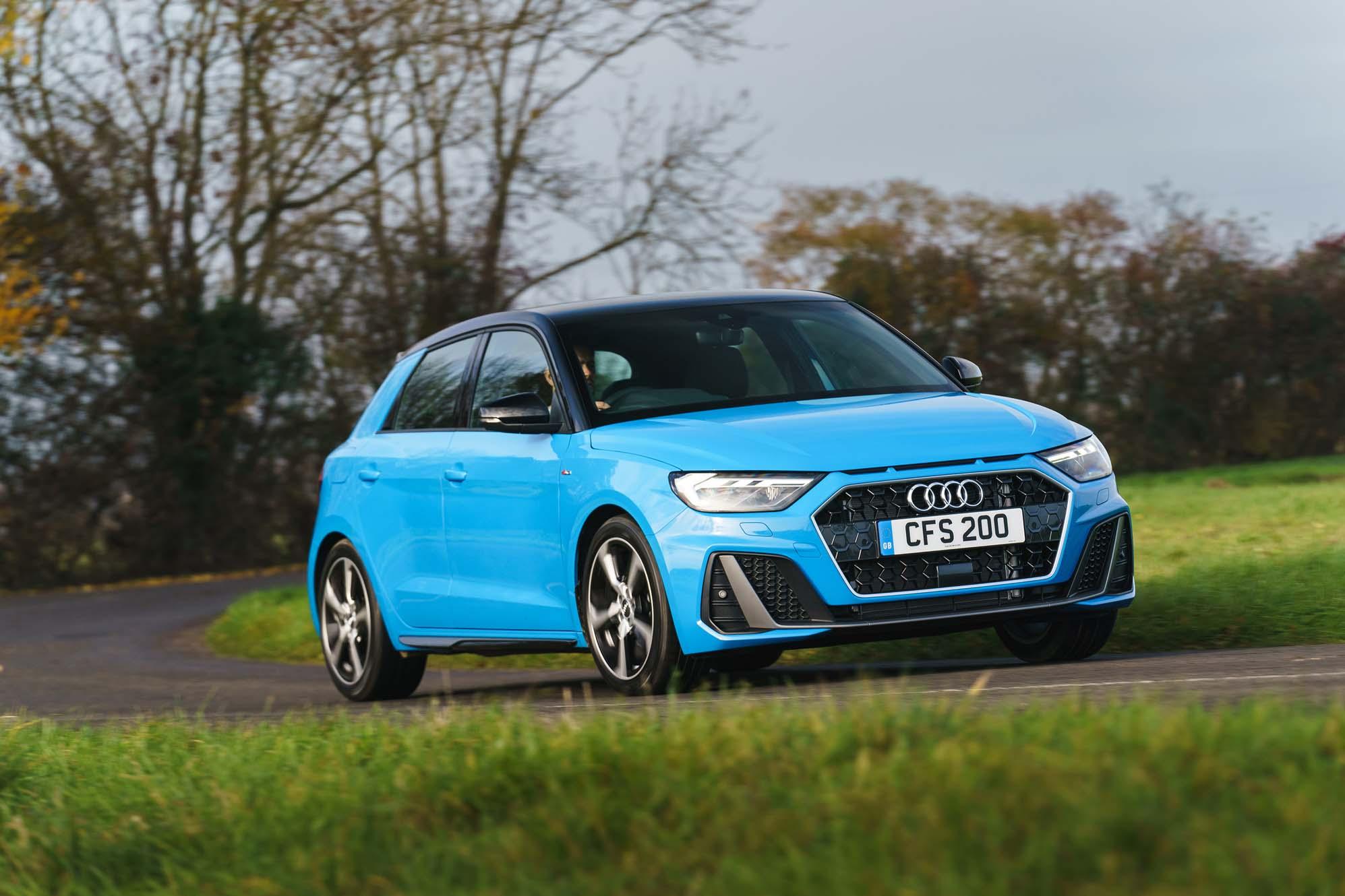 A blue Audi A1 driving on a rural road with green grass and bare trees in the background.