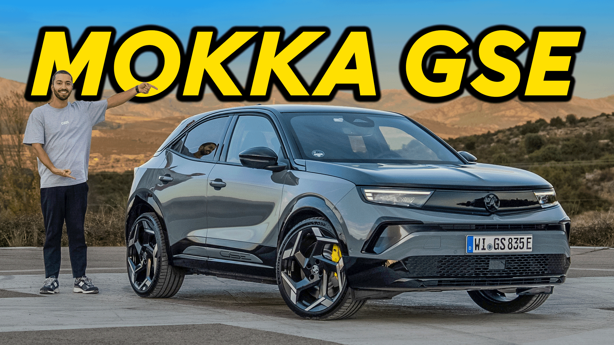 Man pointing at a gray Mokka GSE car parked on a scenic overlook, with "MOKKA GSE" in bold yellow text above.