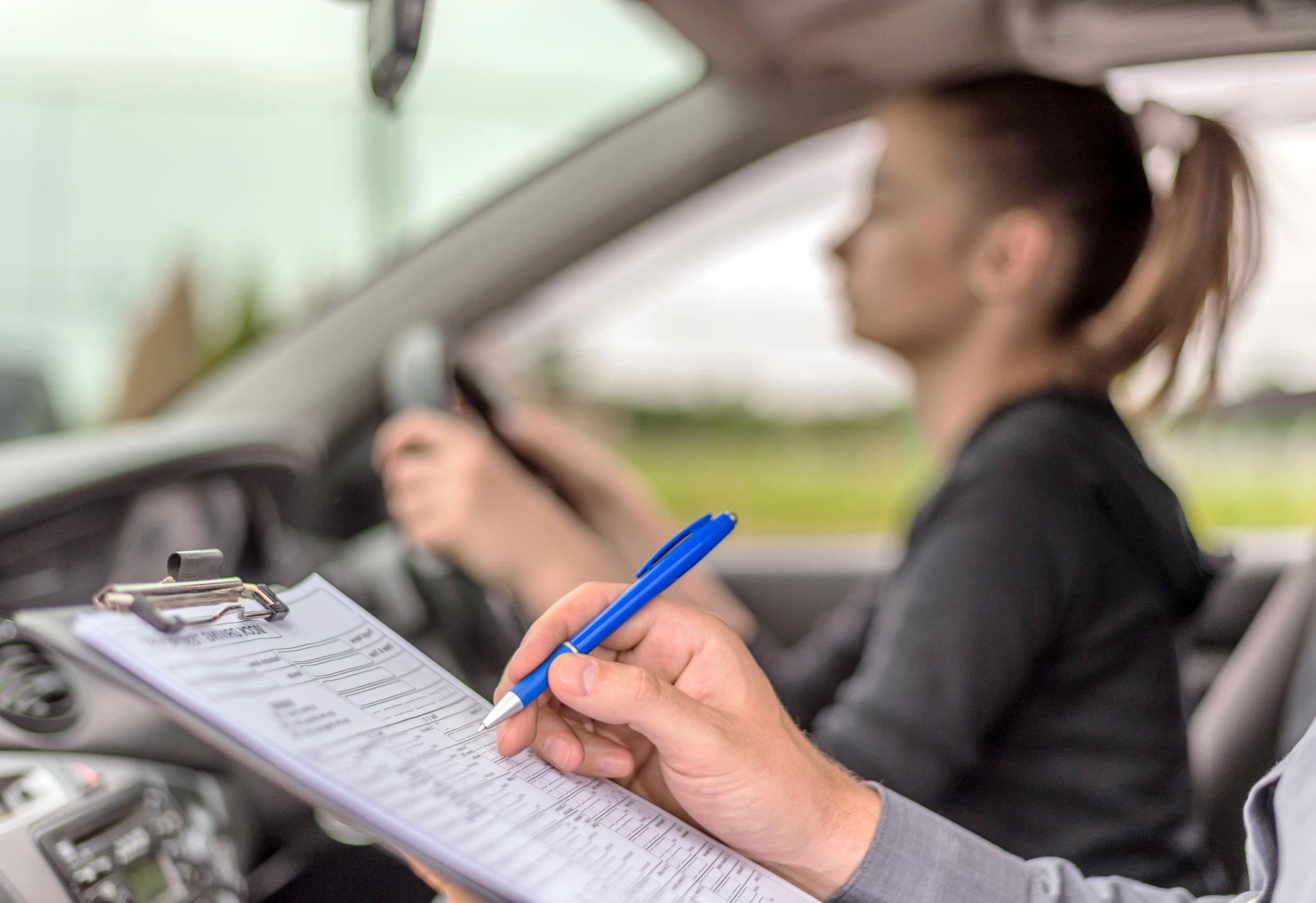 A driving test examiner marking points on a clipboard in the front of a car