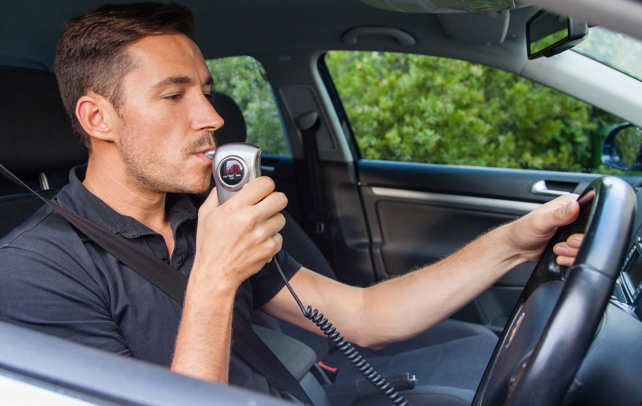 Man in a car using a breathalyser device while holding the steering wheel, with trees visible through the window.