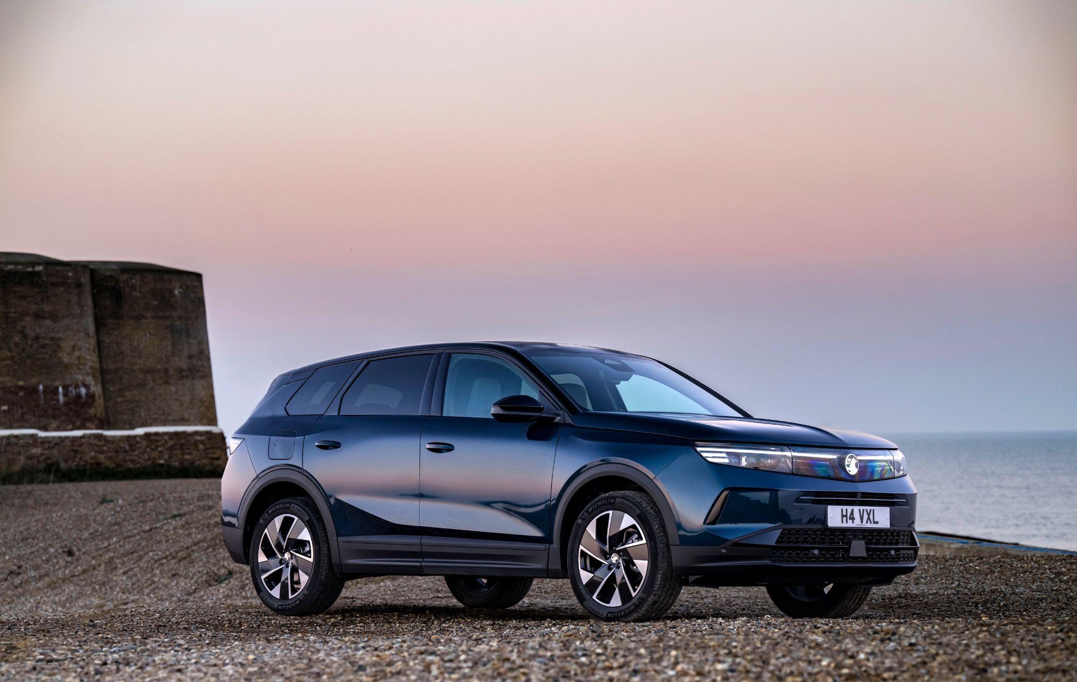 A dark Vauxhall Grandland parked on a gravel surface near a coastal area at sunset, with a calm sea and a clear sky in the background.