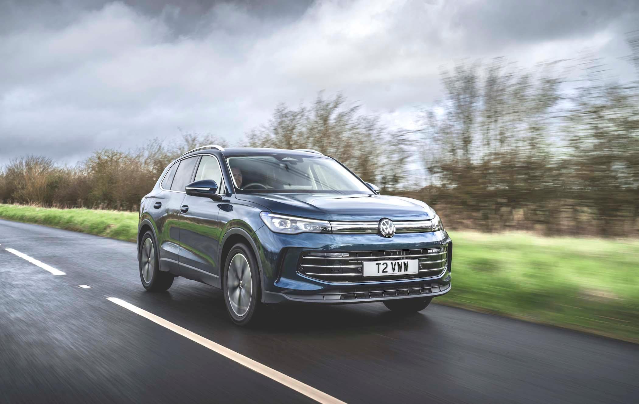 A blue VW Tiguan drives on a rural road with blurred trees in the background under a cloudy sky.