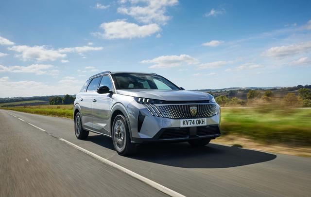 Silver Peugeot 5008 driving on a rural road under a blue sky with scattered clouds, surrounded by green fields and distant hills.