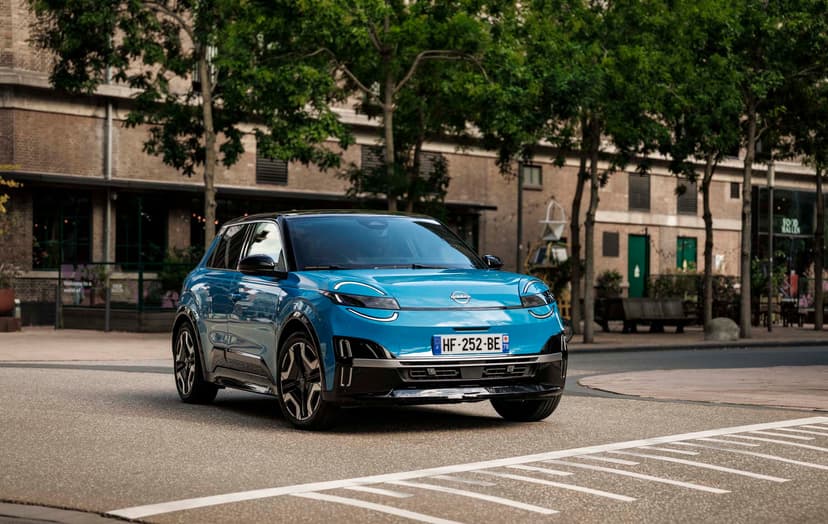 Blue Nissan MICRA driving on a city street, surrounded by green trees and brick buildings.