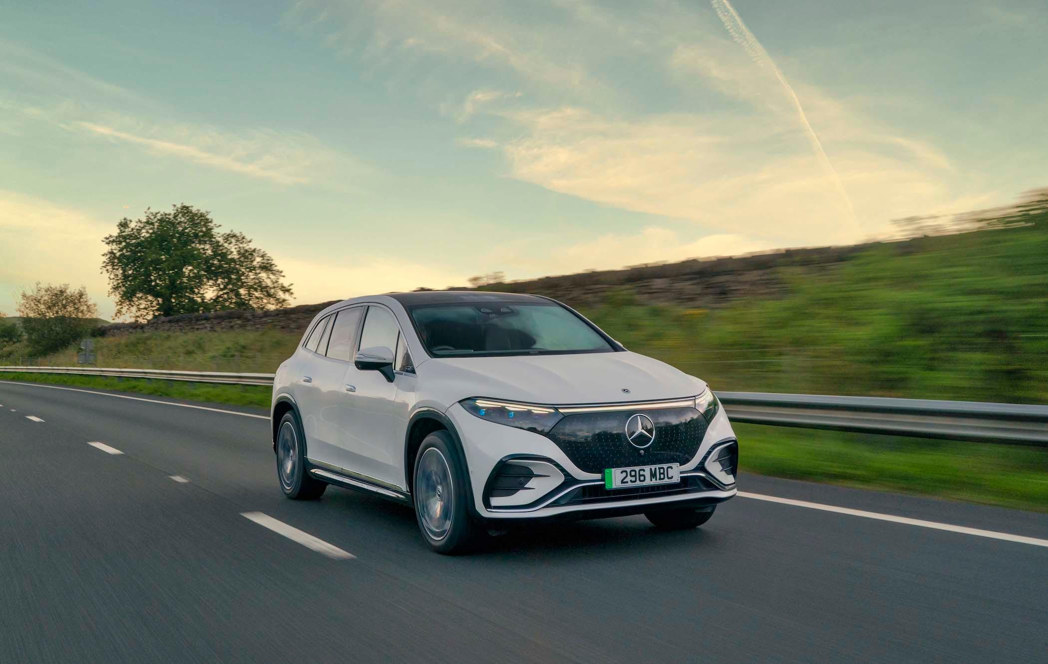 A white Mercedes EQS SUV driving on a highway with a grassy landscape and a tree in the background under a partly cloudy sky.