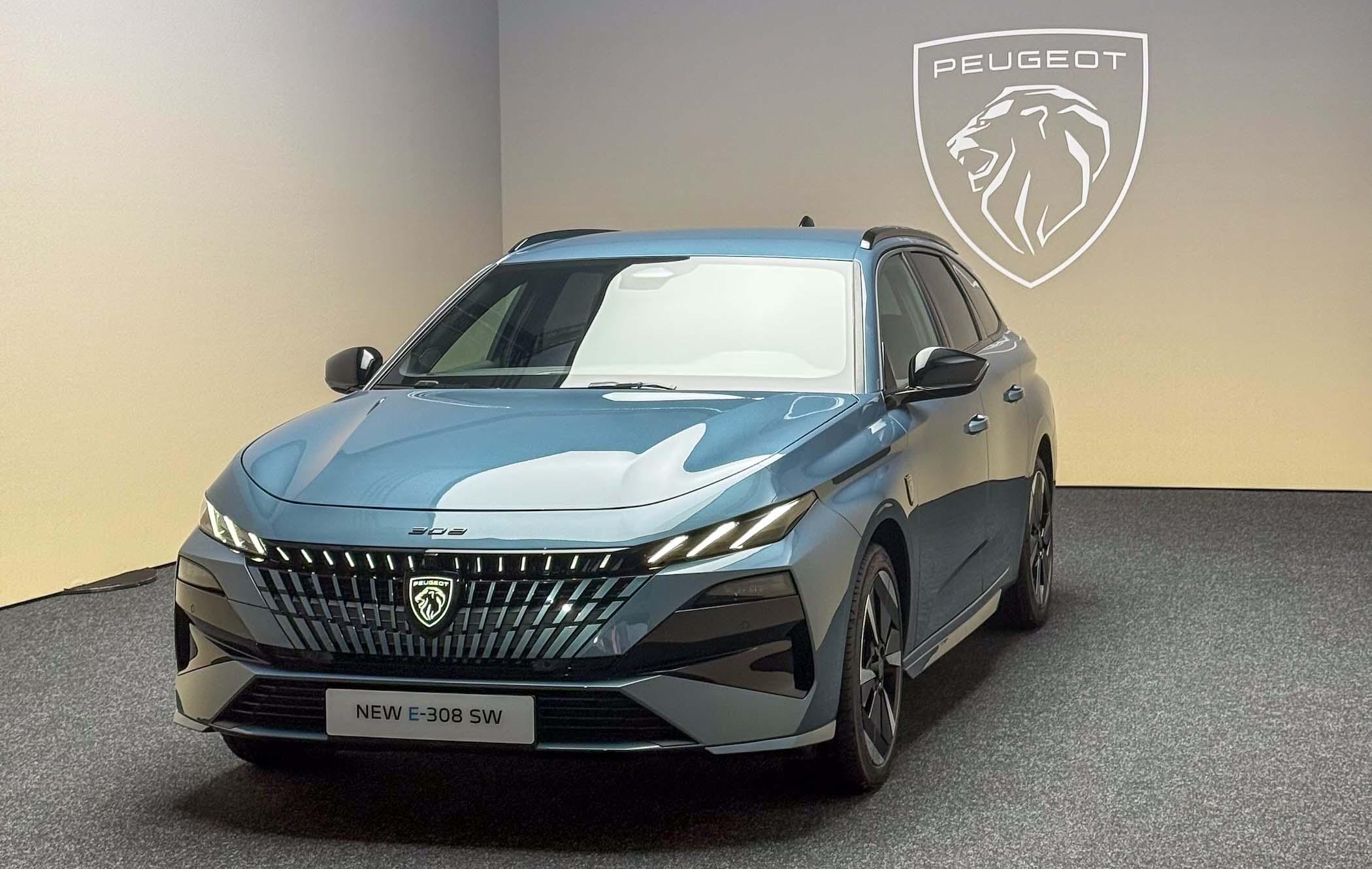Facelifted Ingaro Blue Peugeot e-308 SW parked in a studio with Peugeot emblem illuminated behind it
