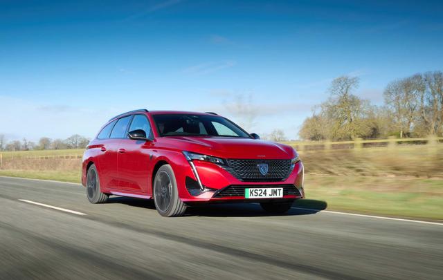 Red Peugeot e-308 SW driving on a rural road with trees and fields in the background under a clear blue sky.