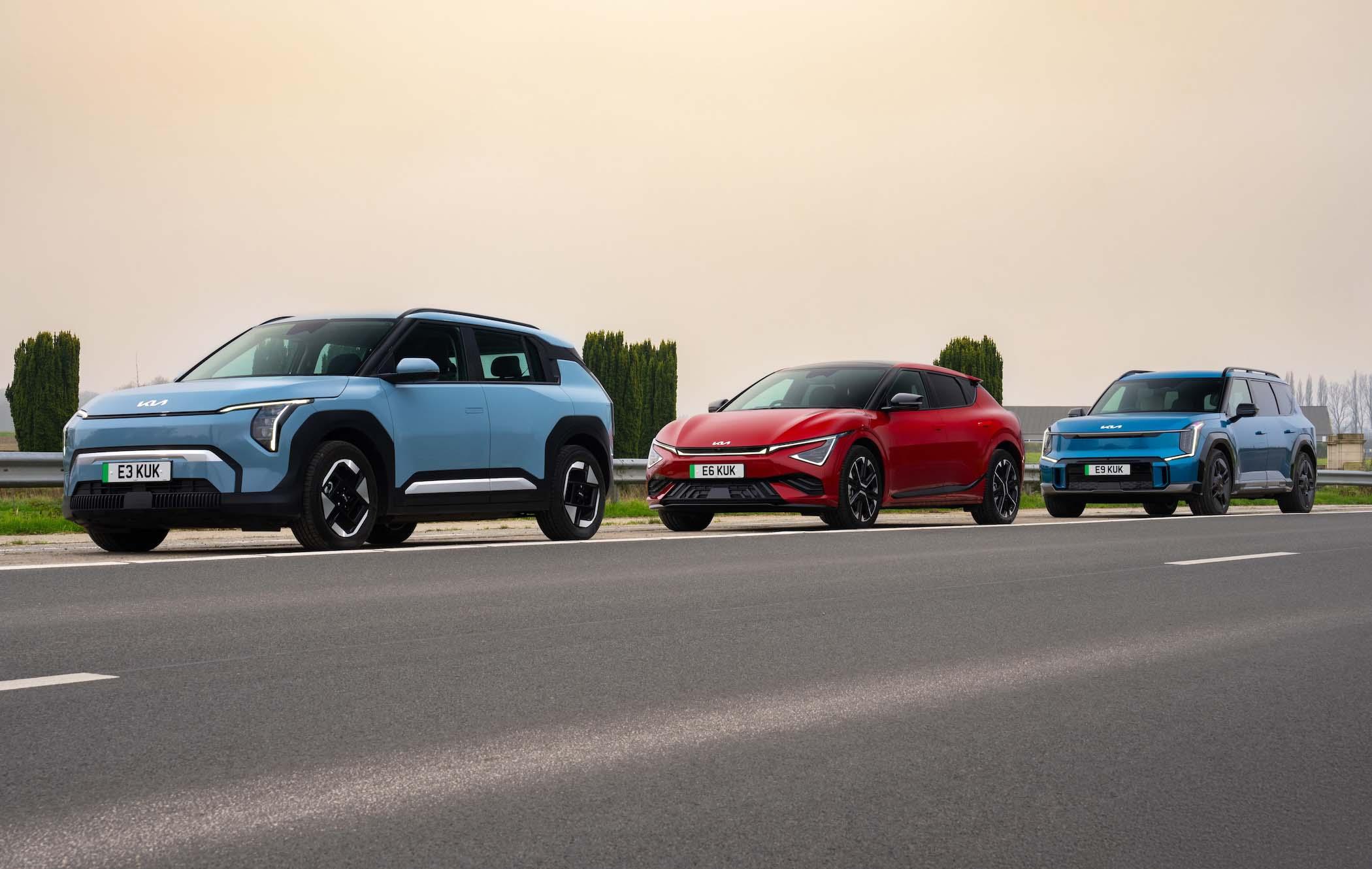 One blue Kia EV3, one red Kia EV6, and a blue Kia EV9 parked in a row on a road with trees in the background.