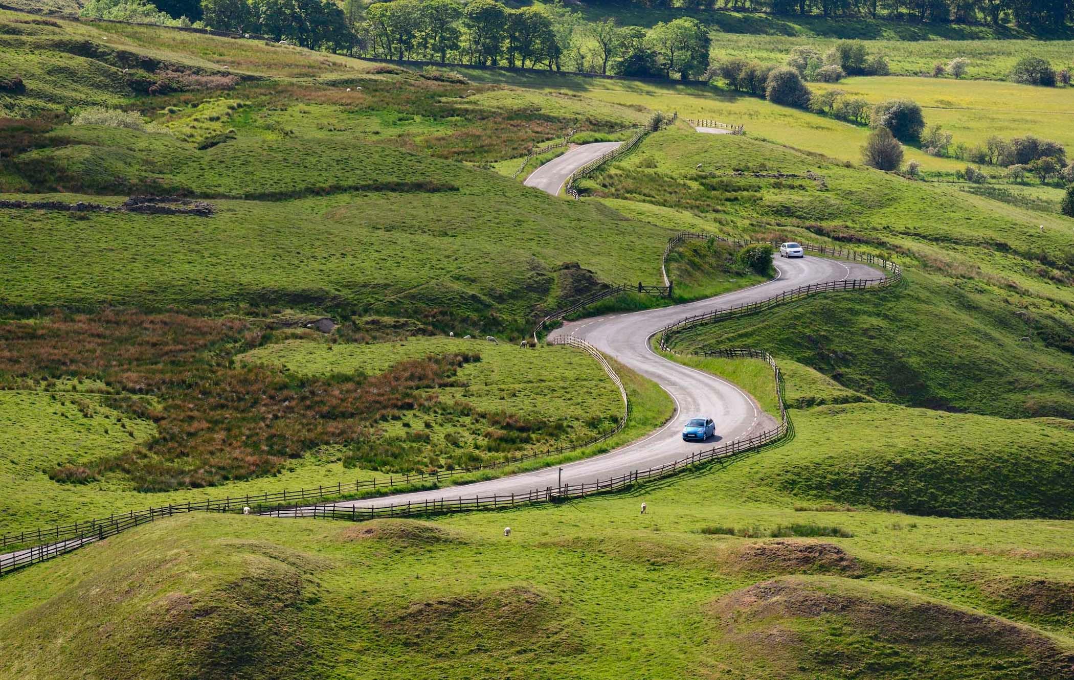 A winding road curves through lush green hills, with a blue car and a white car driving along. Trees line the horizon under a clear sky.