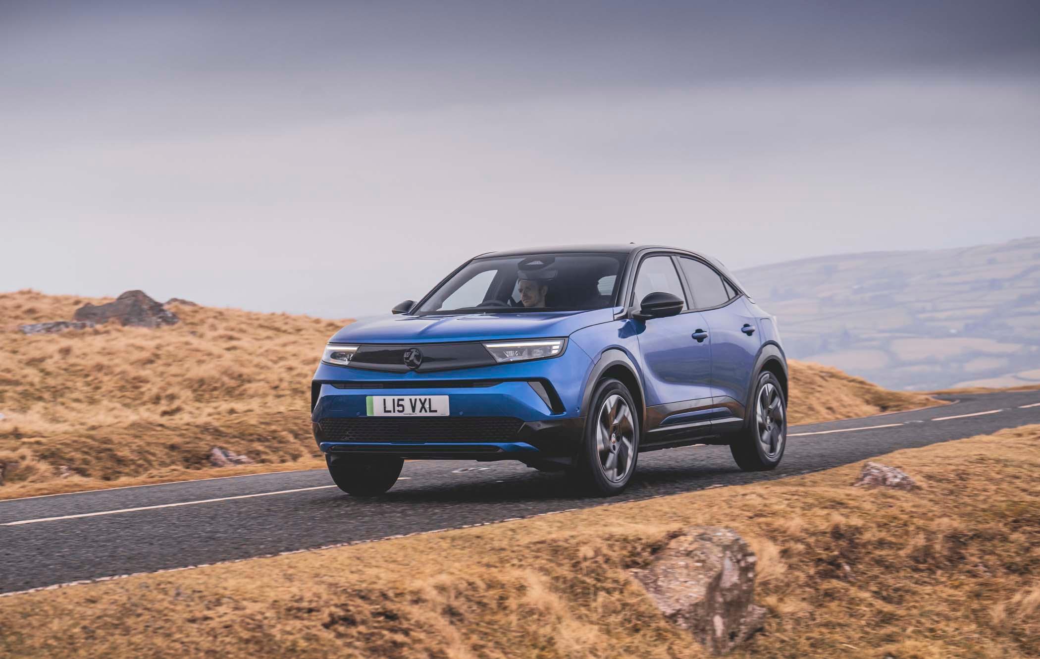 A blue Vauxhall Mokka Electric drives on a rural road through a hilly landscape under a cloudy sky.