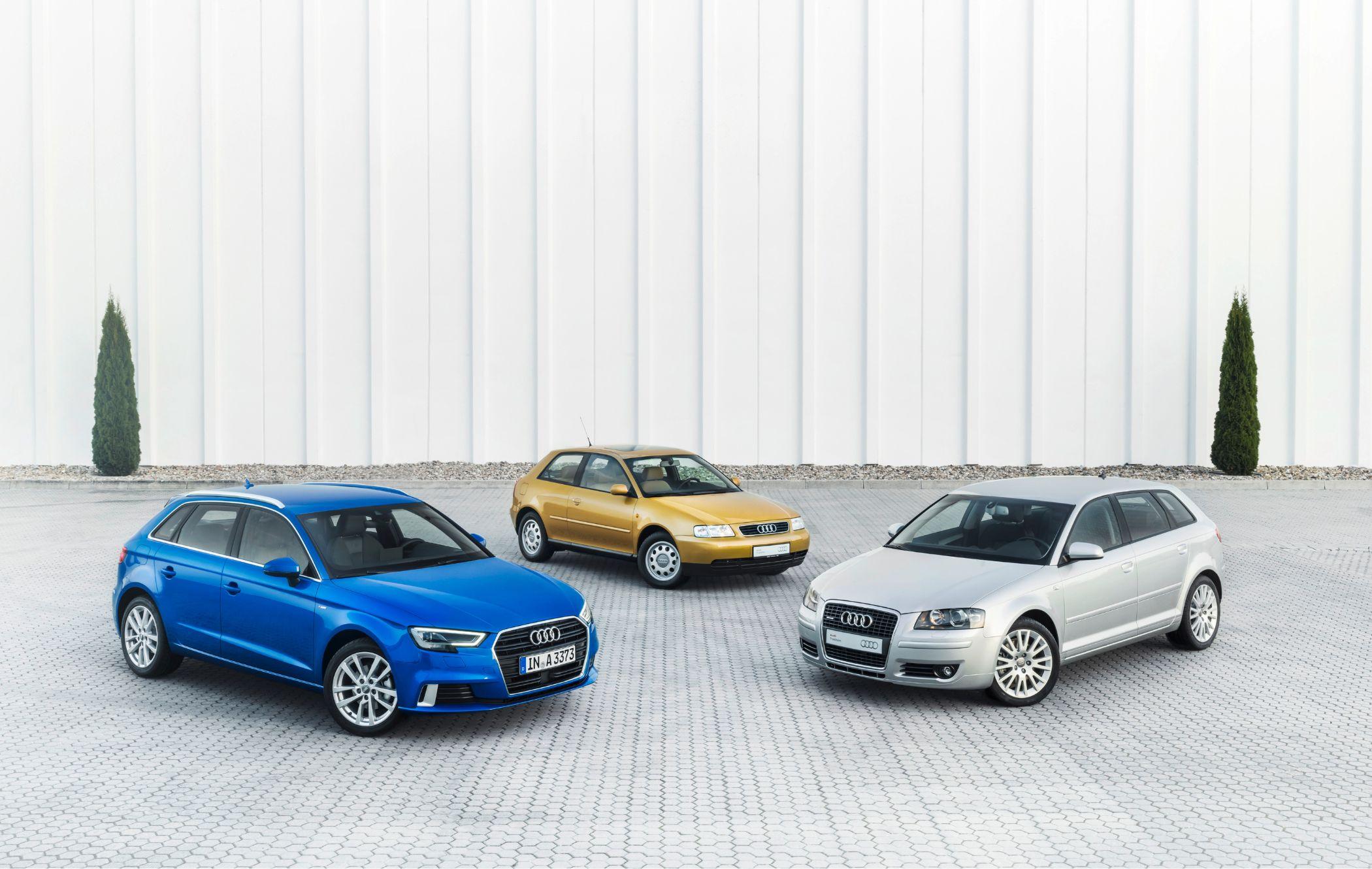 Three Audi A3 hatchbacks in blue, gold, and silver are parked on a paved area with a white wall and two tall shrubs in the background.