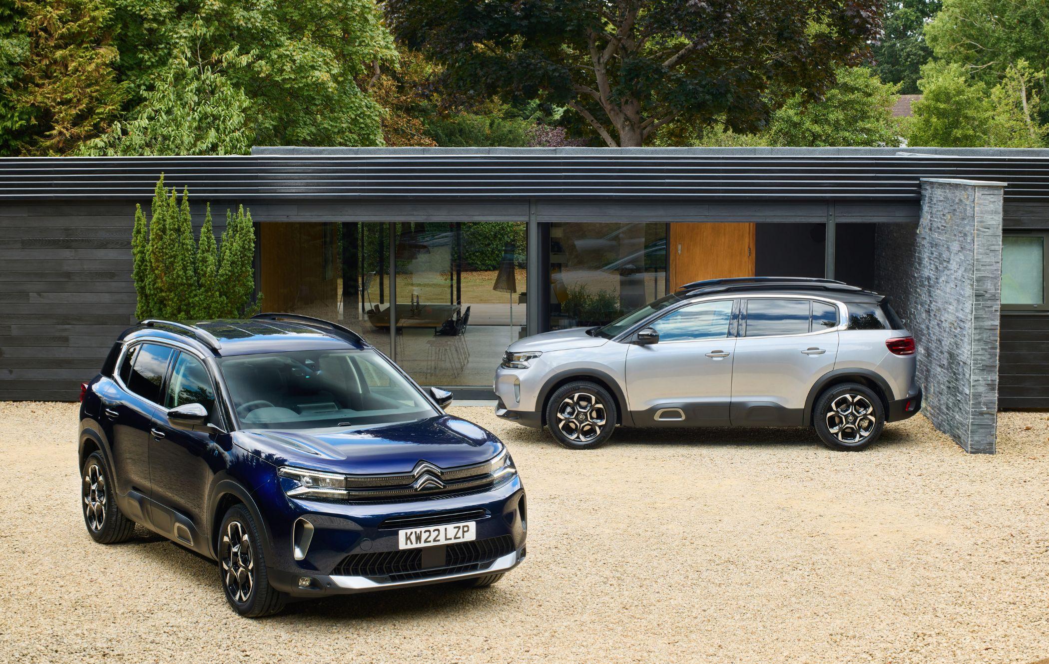 Two Citroën C5 Aircross SUVs parked on a gravel driveway in front of a modern house with large glass windows and surrounded by trees.