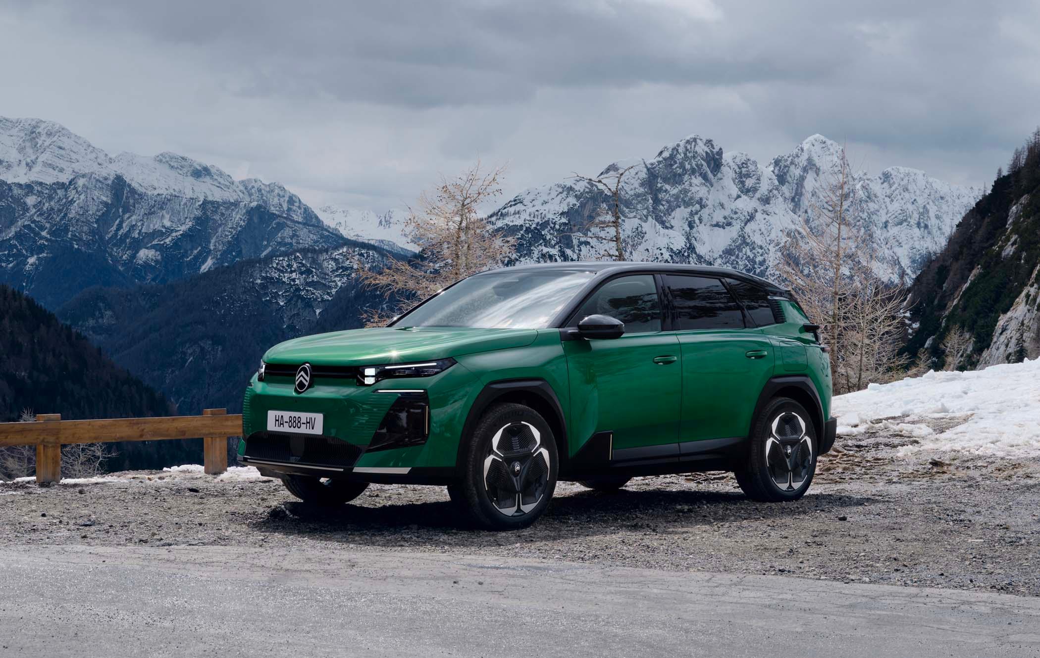 A green Citroën C5 Aircross parked on a mountain road with snow-covered peaks and cloudy skies in the background.