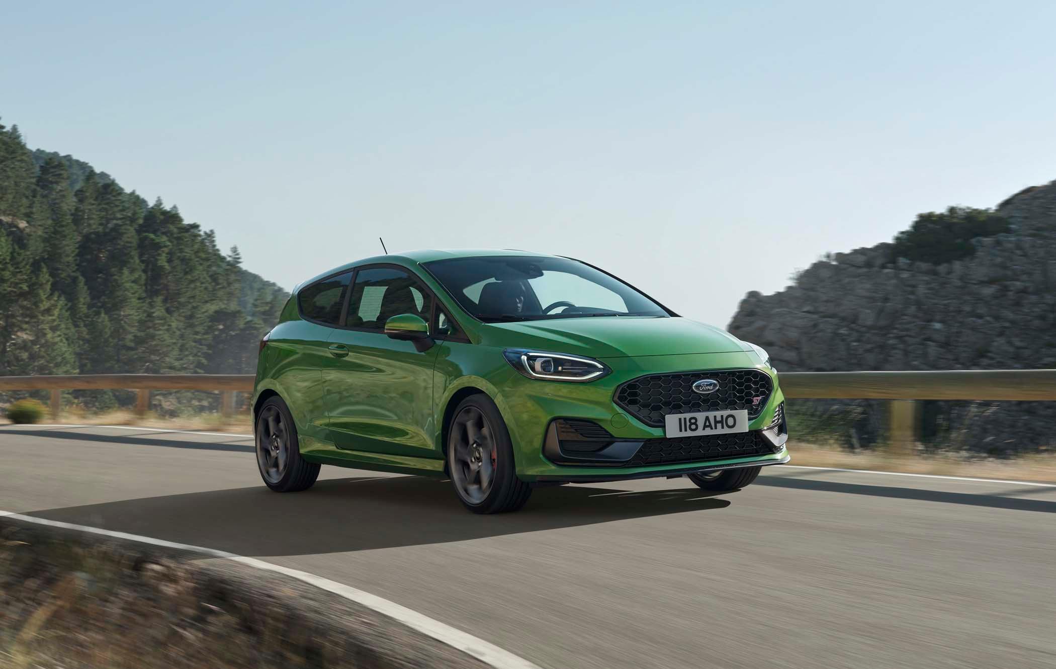 Green Ford Fiesta driving on a scenic mountain road with trees and rocky hills in the background under a clear sky.