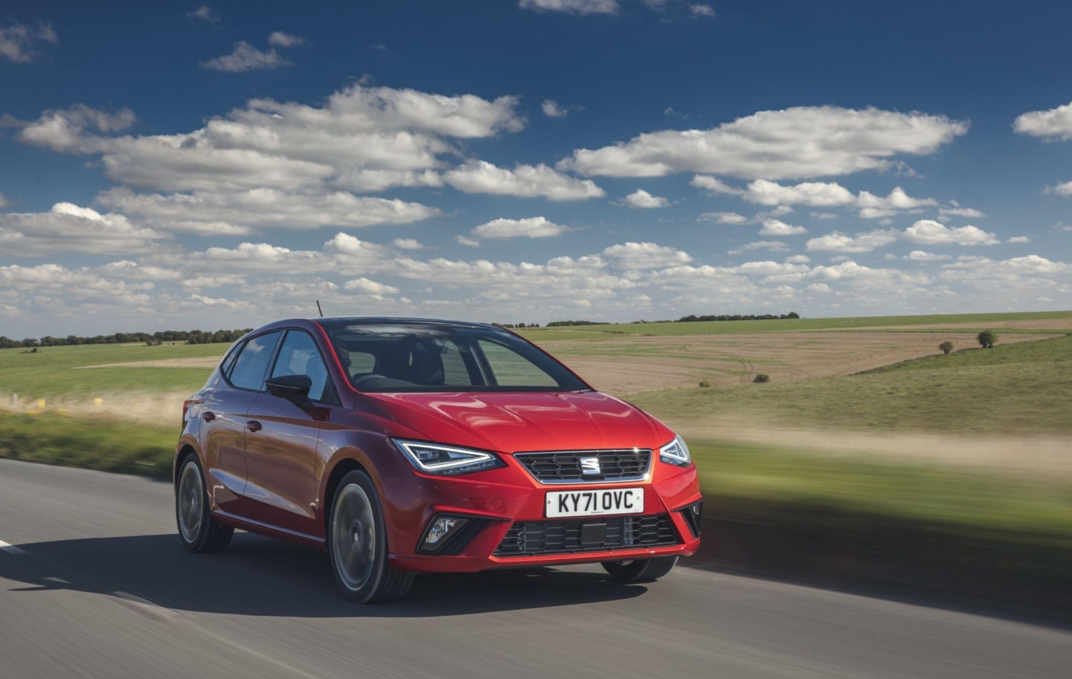 A red hatchback car driving on a countryside road under a blue sky with scattered clouds.