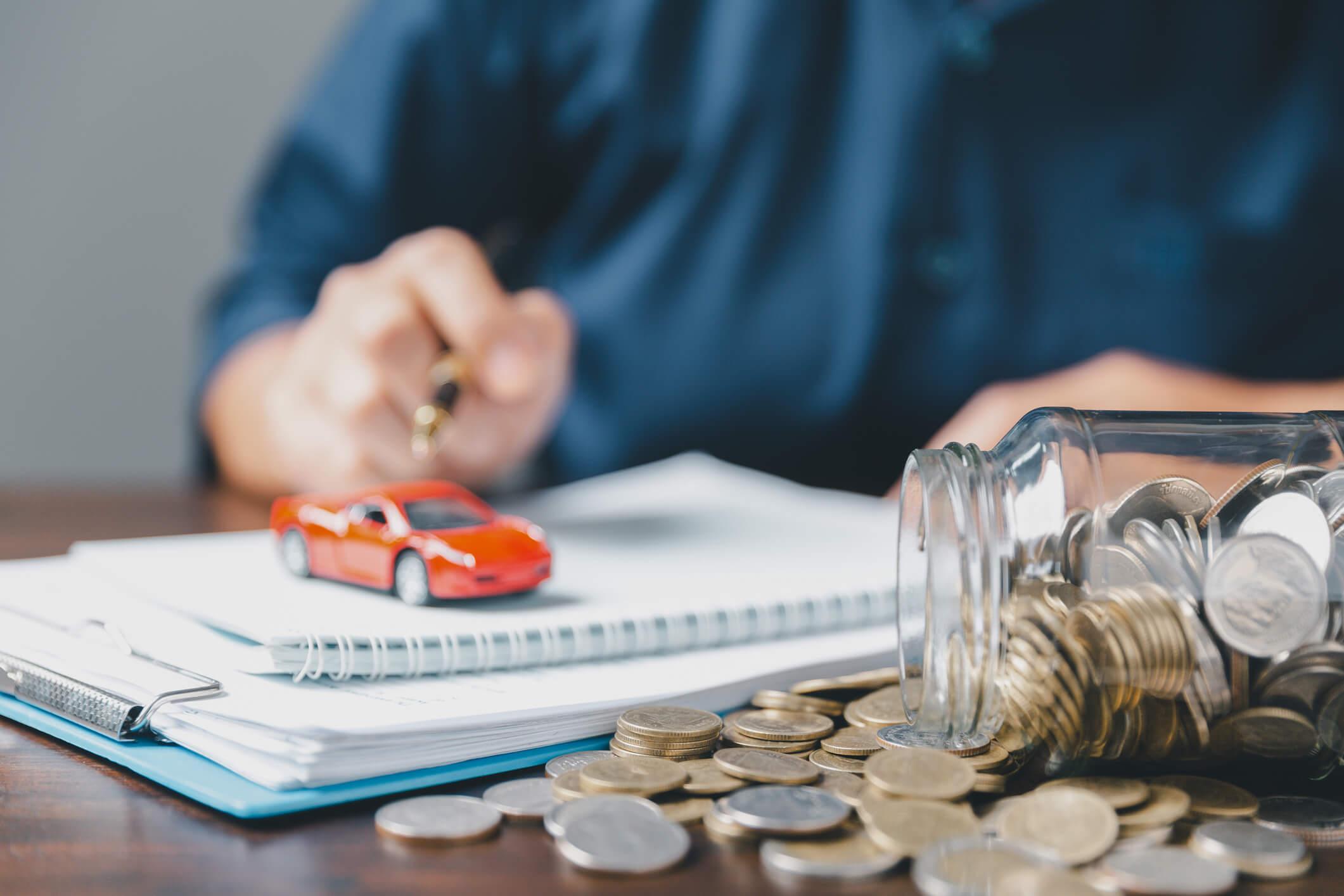 A close up of a desk showing a hand writing and a jar of money, plus a small red toy car