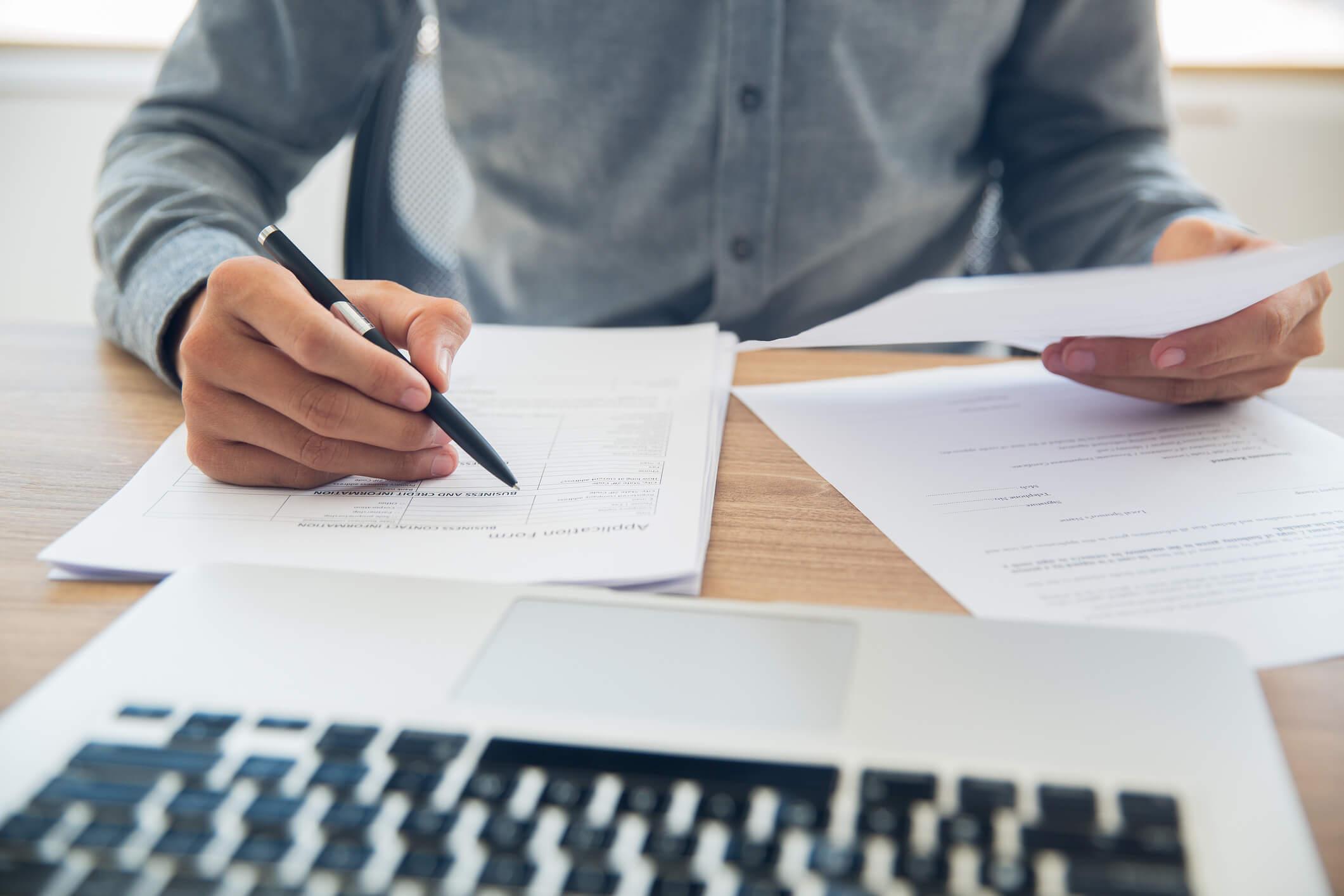 A close up of a laptop on a desk with somebody reviewing documents