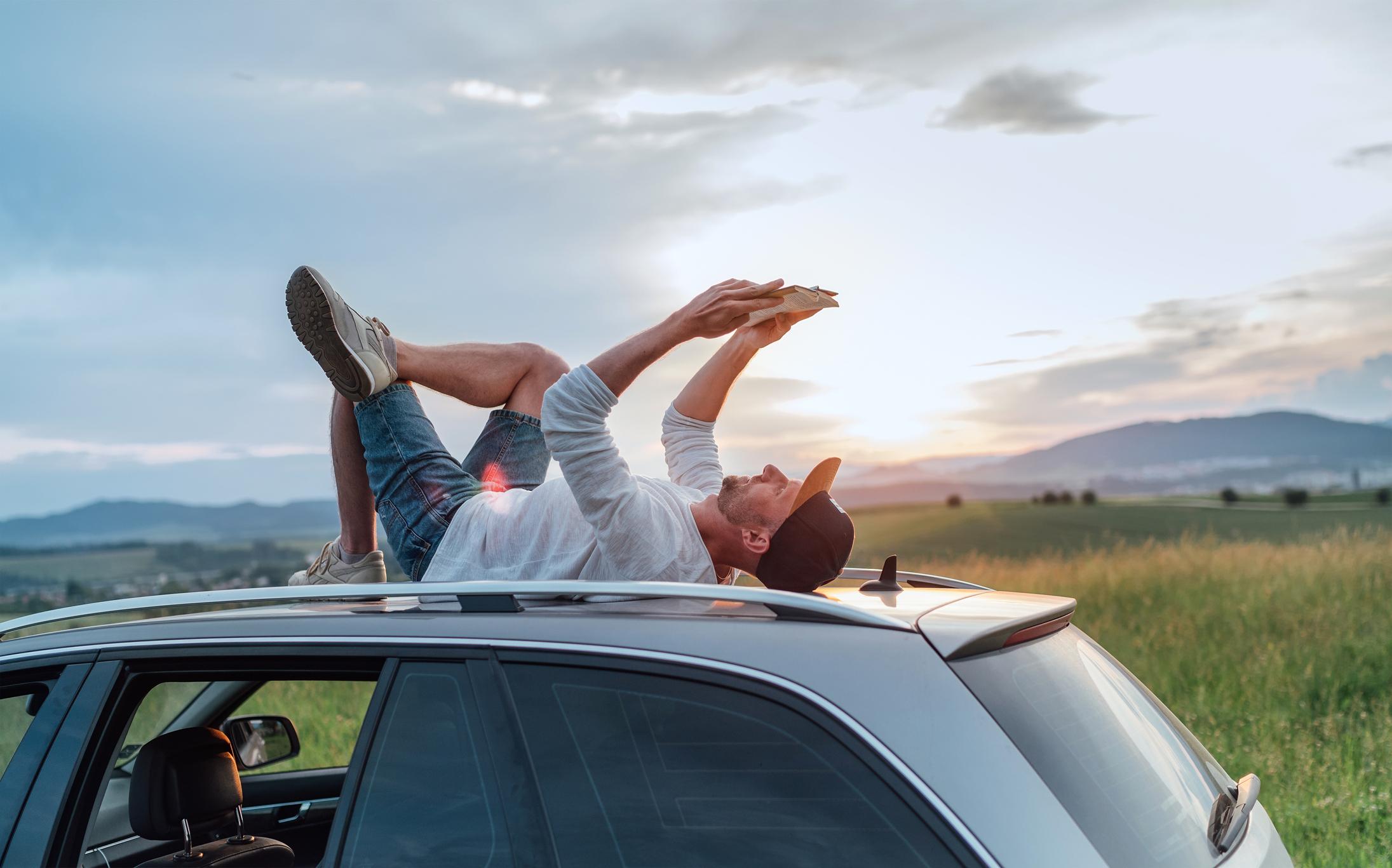 A man laying on the roof of a car reading a book