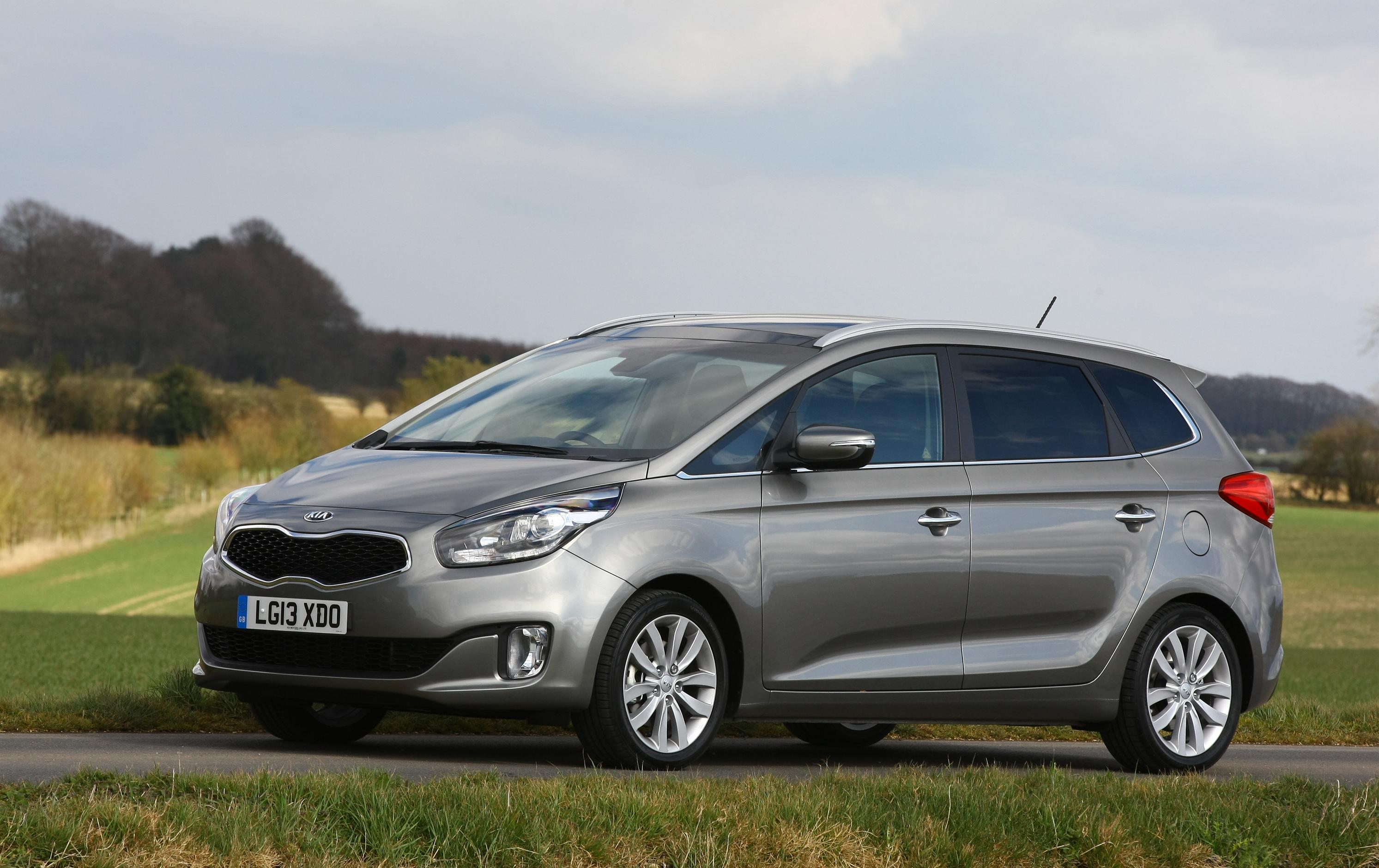 A grey Kia Carens with a field in the background