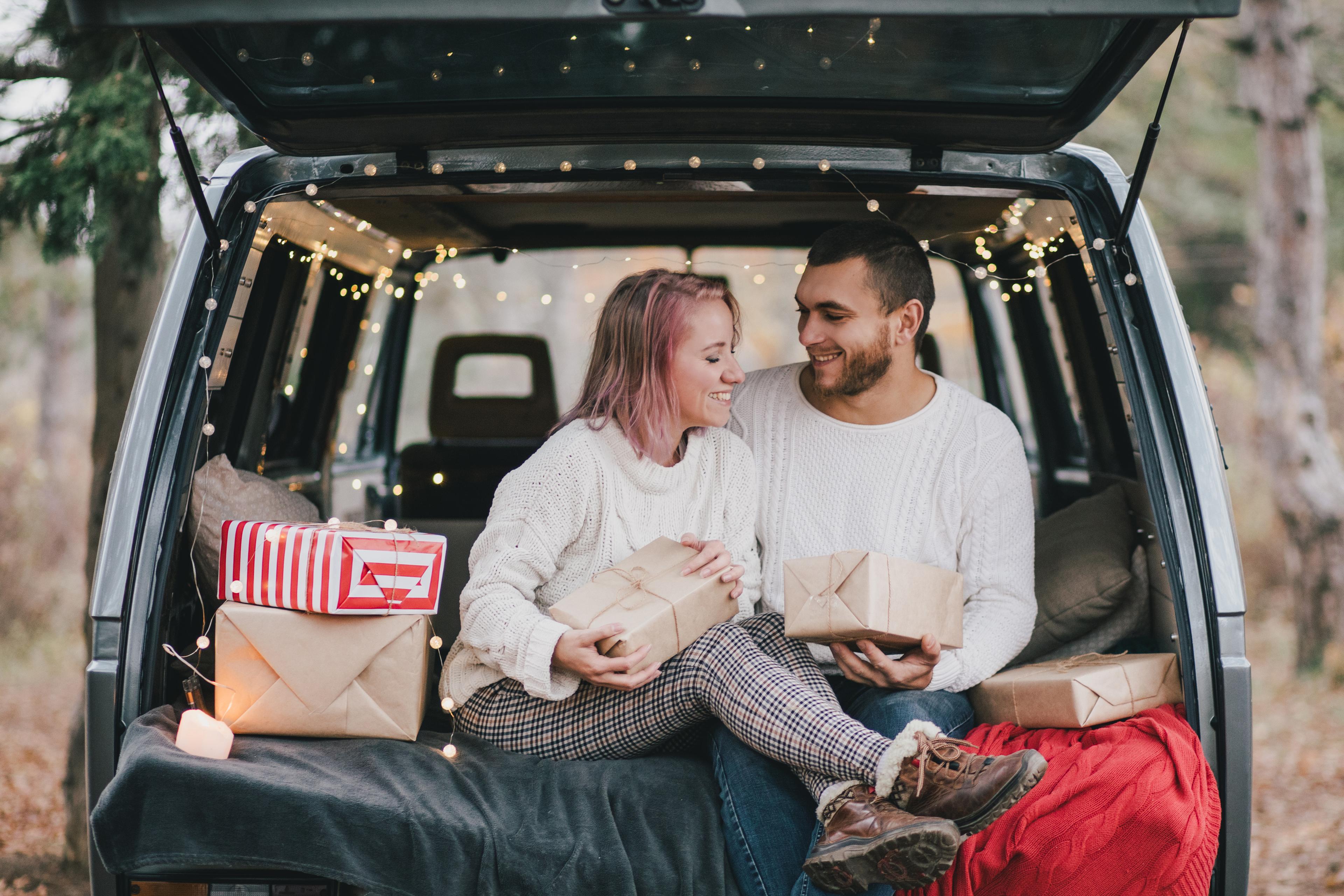 A man and a woman sitting in the back of a car with Christmas decor and presents. 