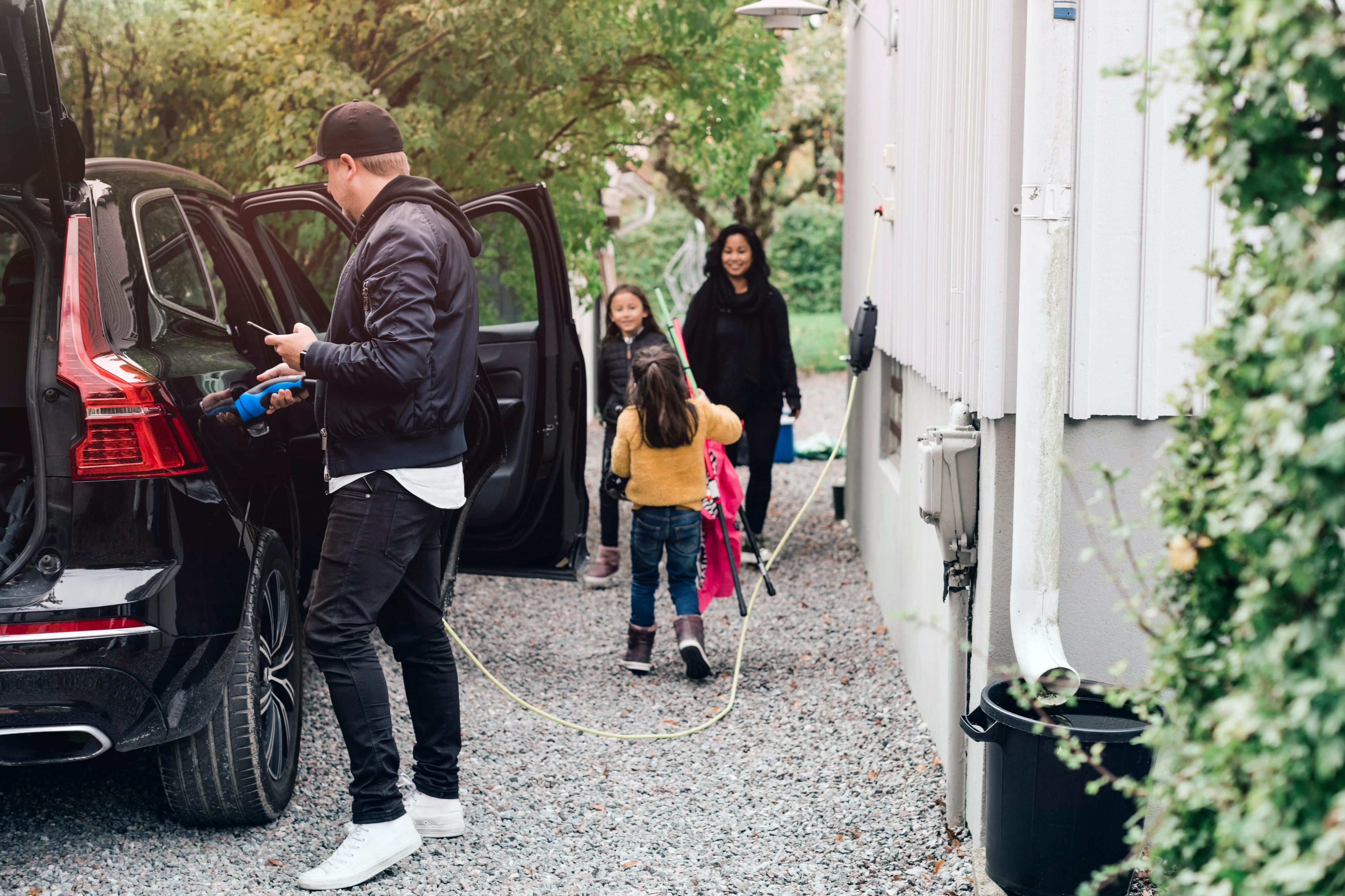 a man charging his black hybrid volvo outside his house while his family gather around