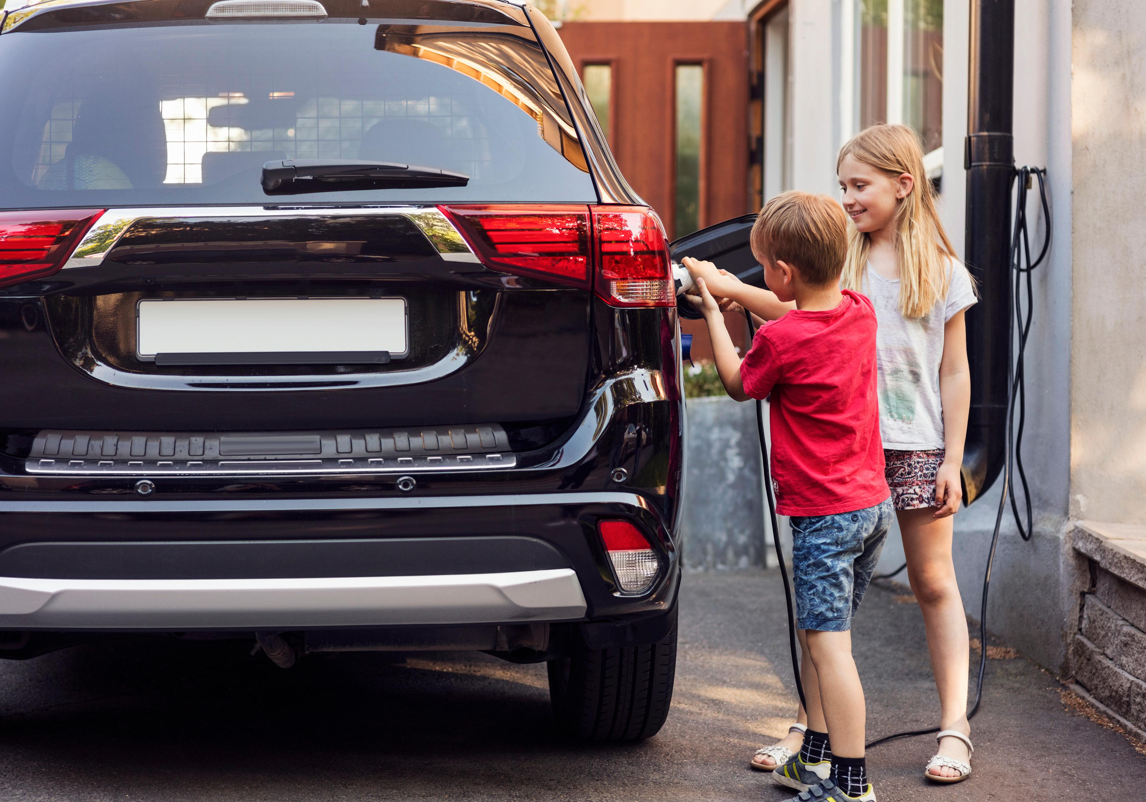 a boy in a red t shirt plugs in a black mitsubishi hybrid to charge, alongside his sister