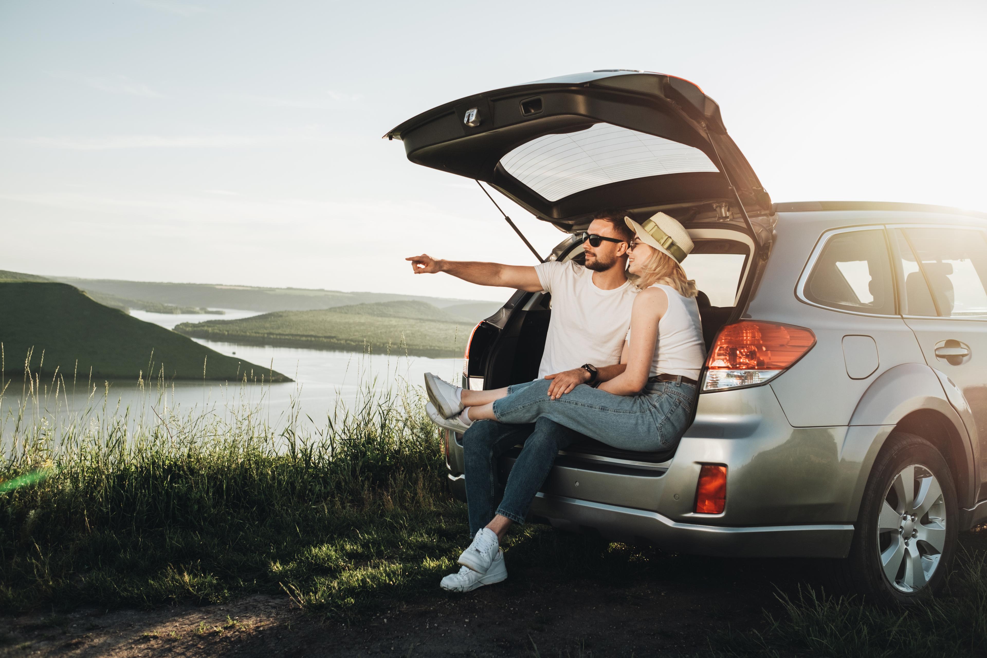 Two people say in the back of a car with the boot open. One is pointing at the mountains and water in the distance