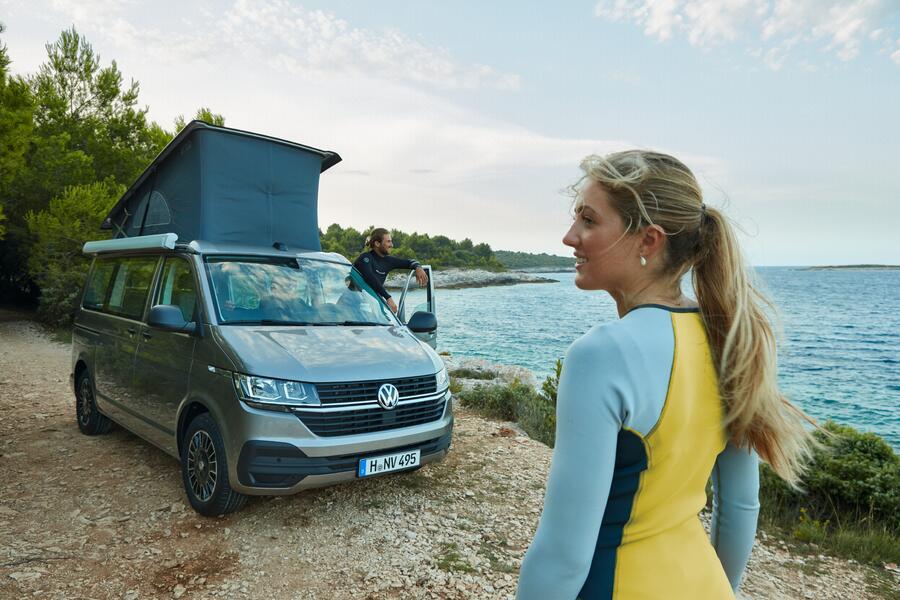 a grey volkswagen california campervan parked by the water next to a girl in a yellow wet suit