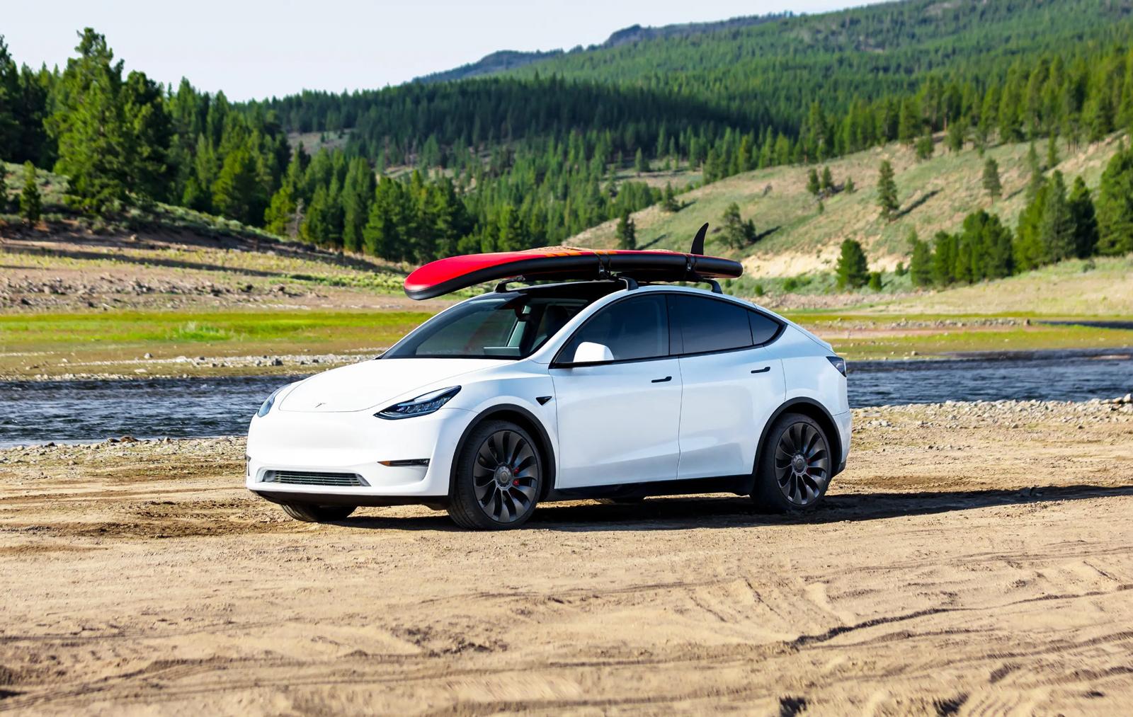 a white Tesla Model Y parked on the short with a red surfboard on the roof