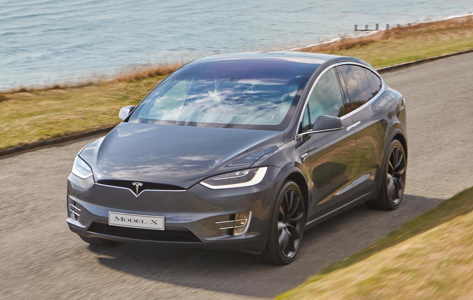 a grey tesla model x driving on a road next to the water
