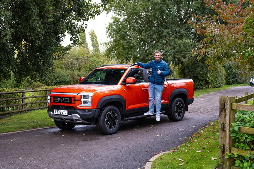 A person in a blue jacket stands beside an orange pickup truck in a lush, leafy countryside setting, giving a thumbs up.