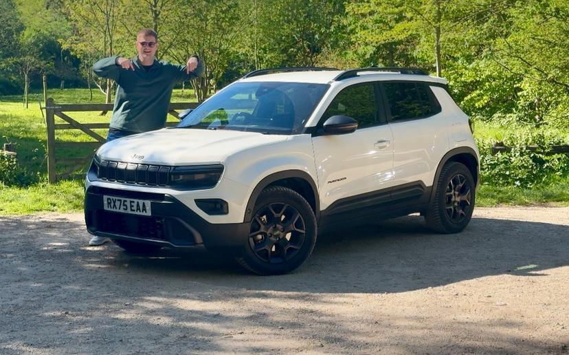 Man pointing at a white SUV parked on a dirt path, surrounded by green trees.