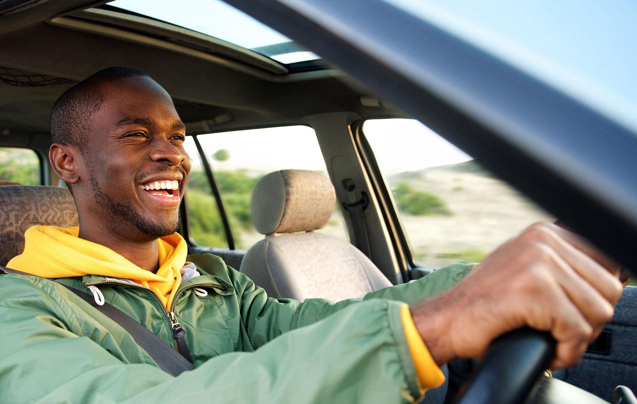 A smiling man sits in his car with his hands on the steering wheel