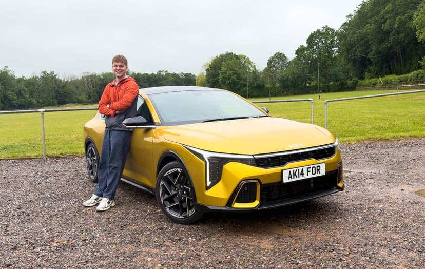 Craig Hale with a Kia K4 parked on a gravel surface, with lush green trees in the background.
