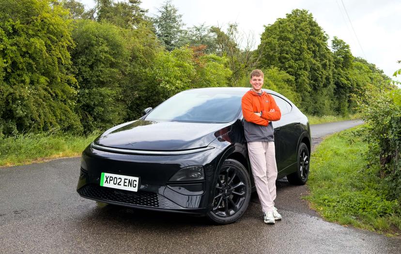 Craig Hale stands beside a black XPENG G6 on a wet road surrounded by lush green trees, wearing an orange jacket and smiling.