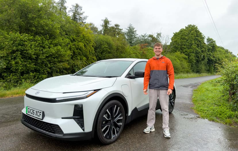 Craig Hale stands beside a white car on a rural road, surrounded by lush green trees under a cloudy sky.