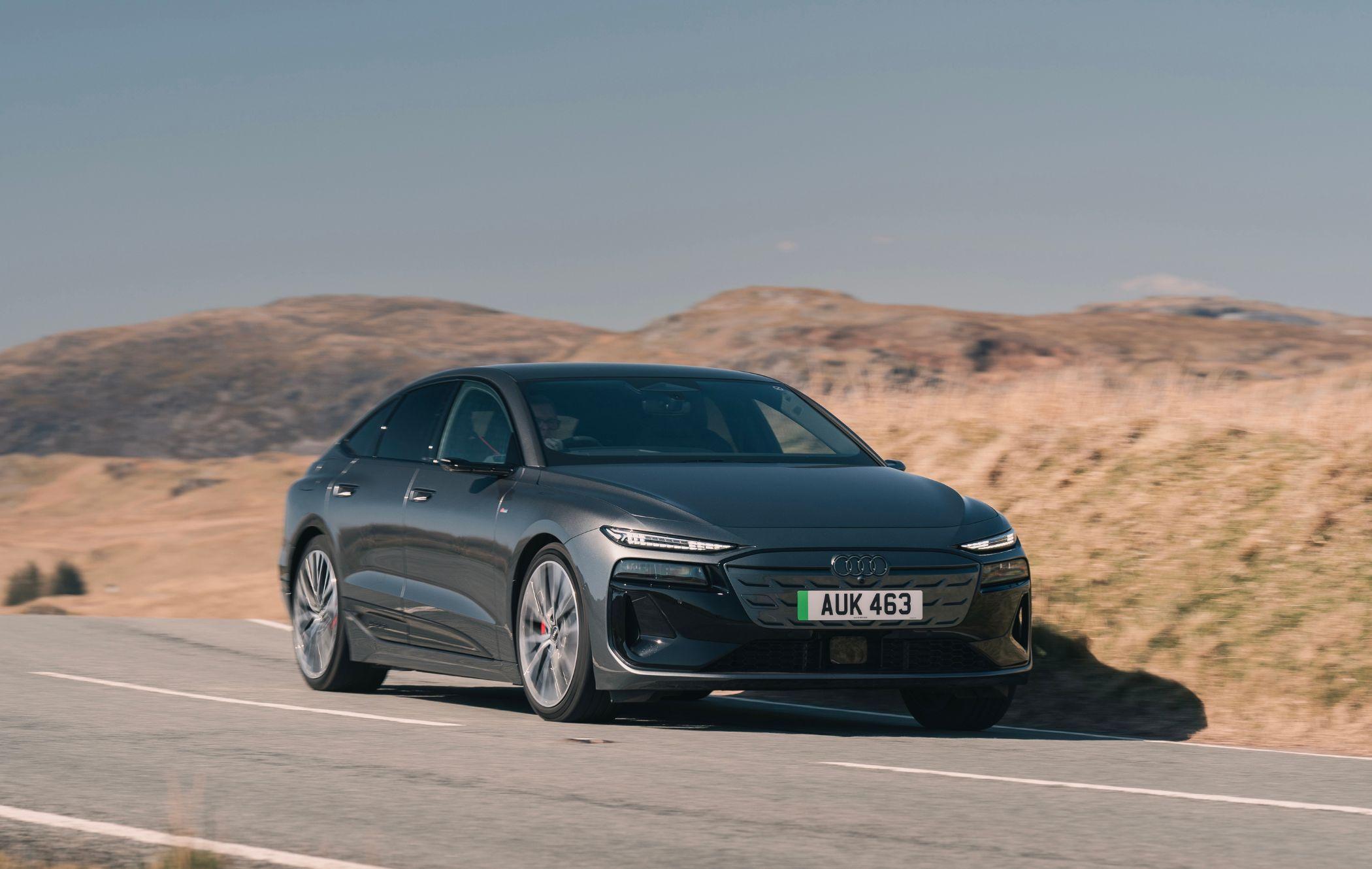 A black Audi A6 e-tron driving on a scenic road with rolling hills in the background under a clear blue sky.