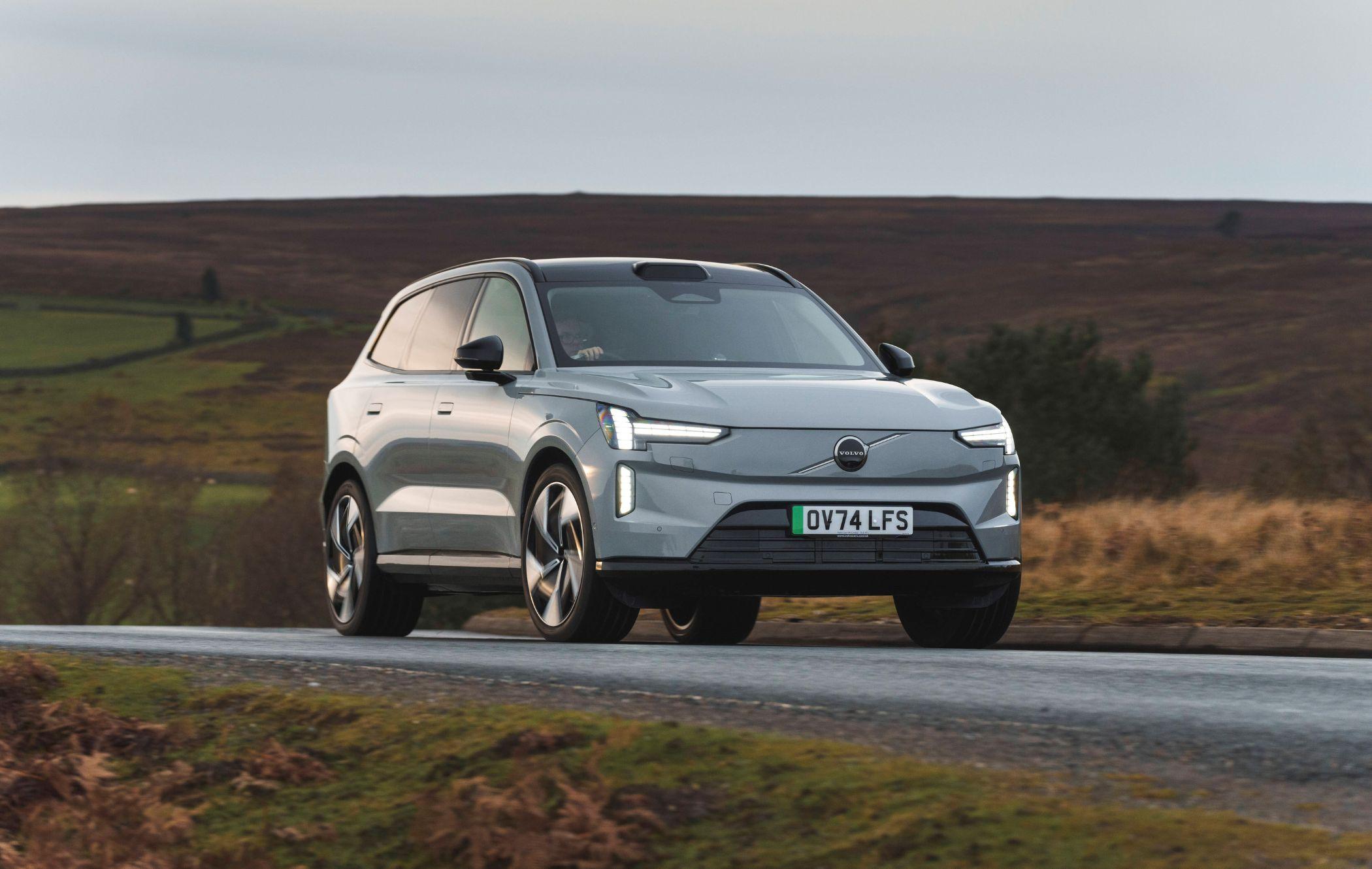 Silver Volvo XC90 driving on a rural road with a grassy landscape and overcast sky in the background.