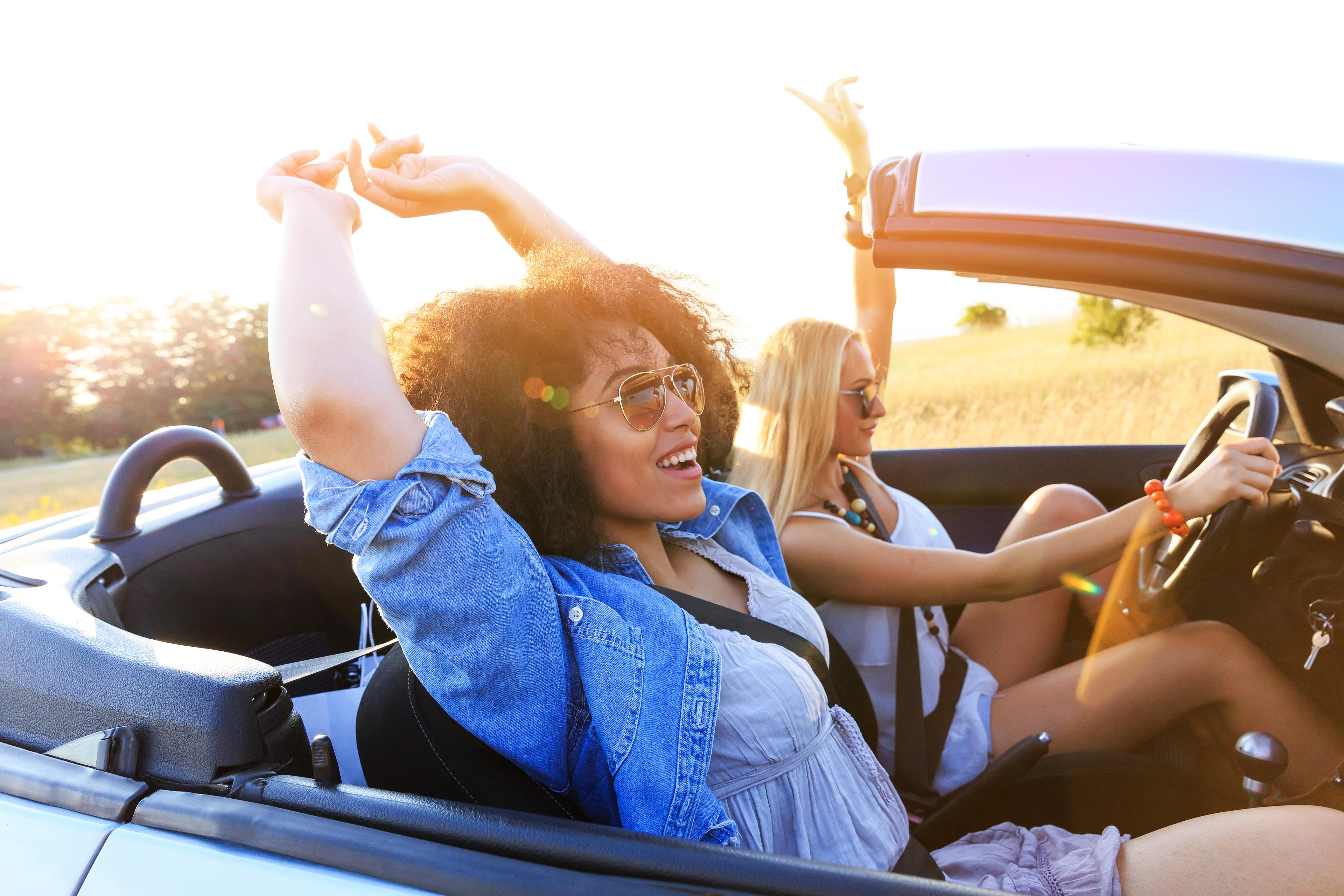 Two women sat in a convertible car with their arms in the air, enjoying the sun. 