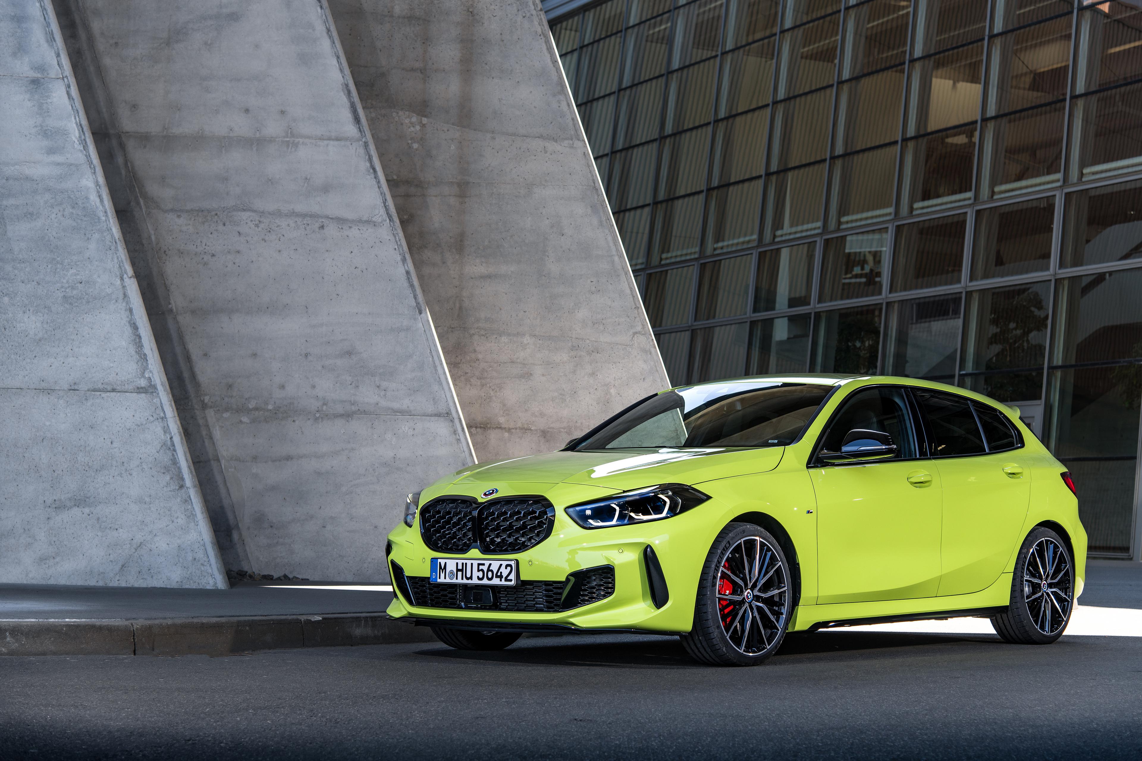 A yellow BMW 1 Series M135i model parked next to a large stone and glass building